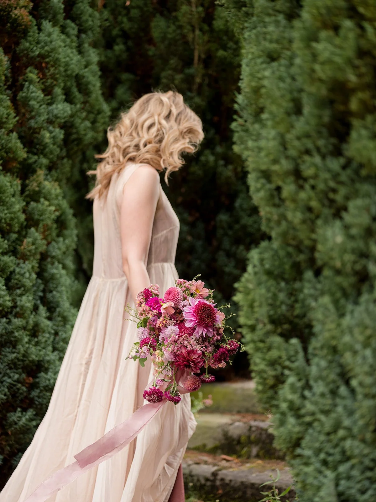 A bride in a cream-colored dress holding a large bouquet of pink and purple flowers, standing between green shrubs in an outdoor setting.
