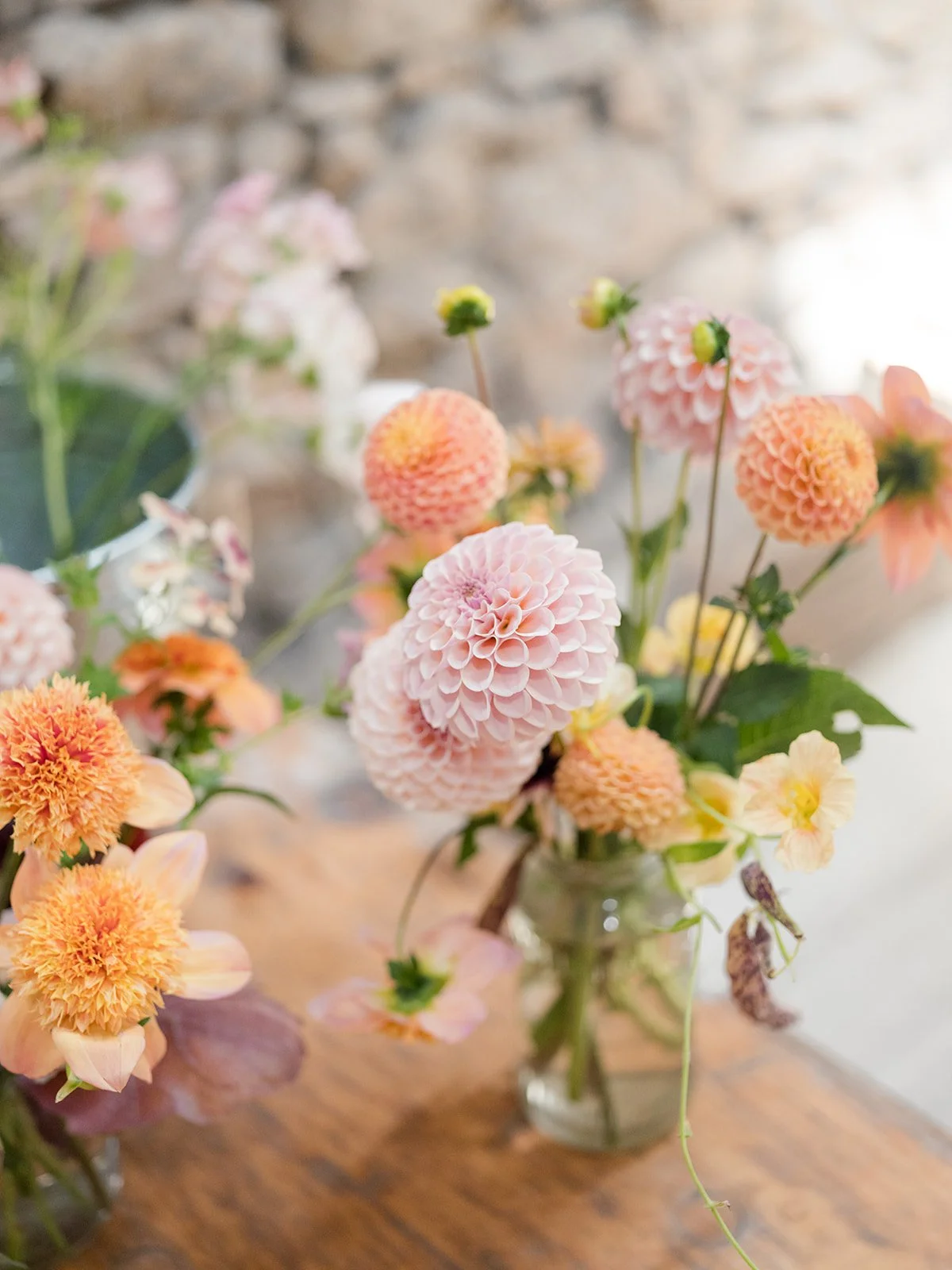 A bouquet of pink, orange, and yellow dahlias in a clear glass vase on a wooden table with a blurred background.