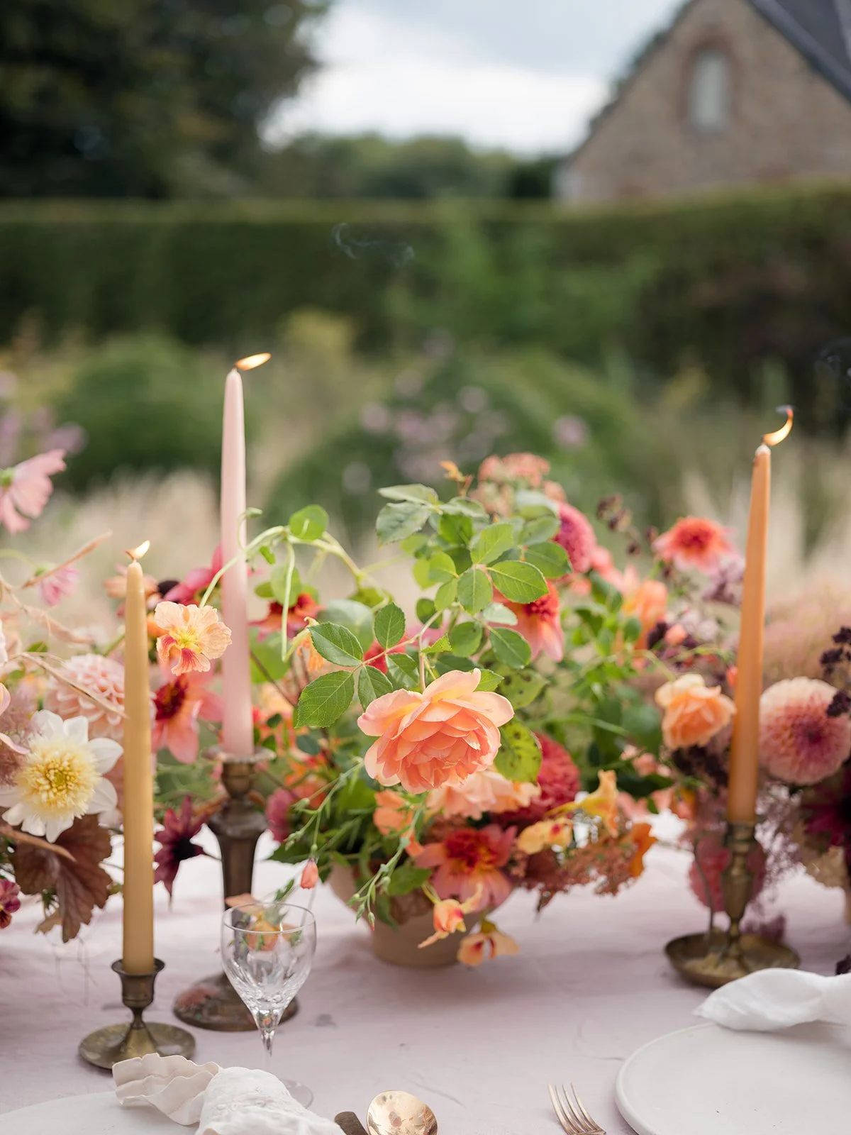 A table centerpiece with pink and peach flowers, candles, and greenery set outdoors with a house and trees in the background.