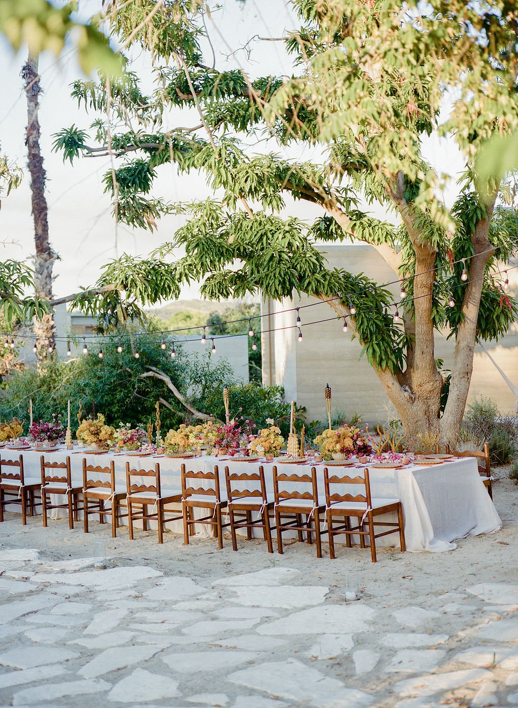 An outdoor dining setup with a long table covered with a white tablecloth, decorated with floral arrangements, set under a large tree with string lights hanging from its branches, on a stone-paved area.