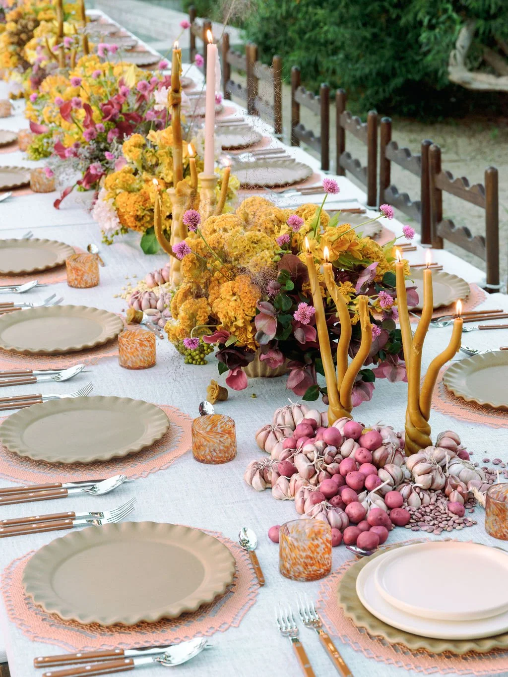 Elegant outdoor dining table decorated with floral arrangements, candles, garlic, and pink hard-boiled eggs, set with plates, cutlery, and glassware.