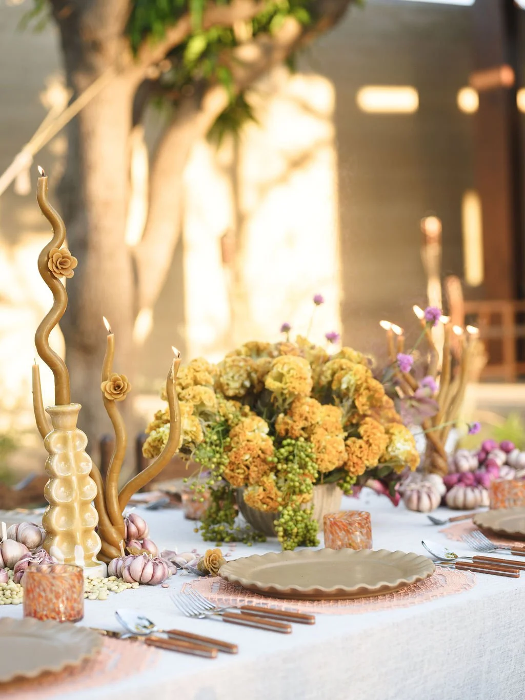 A decorated outdoor dining table with a large floral centerpiece, cream-colored candles, and tableware. The setting is elegant, with soft lighting and a background of trees and blurred outdoor scenery.