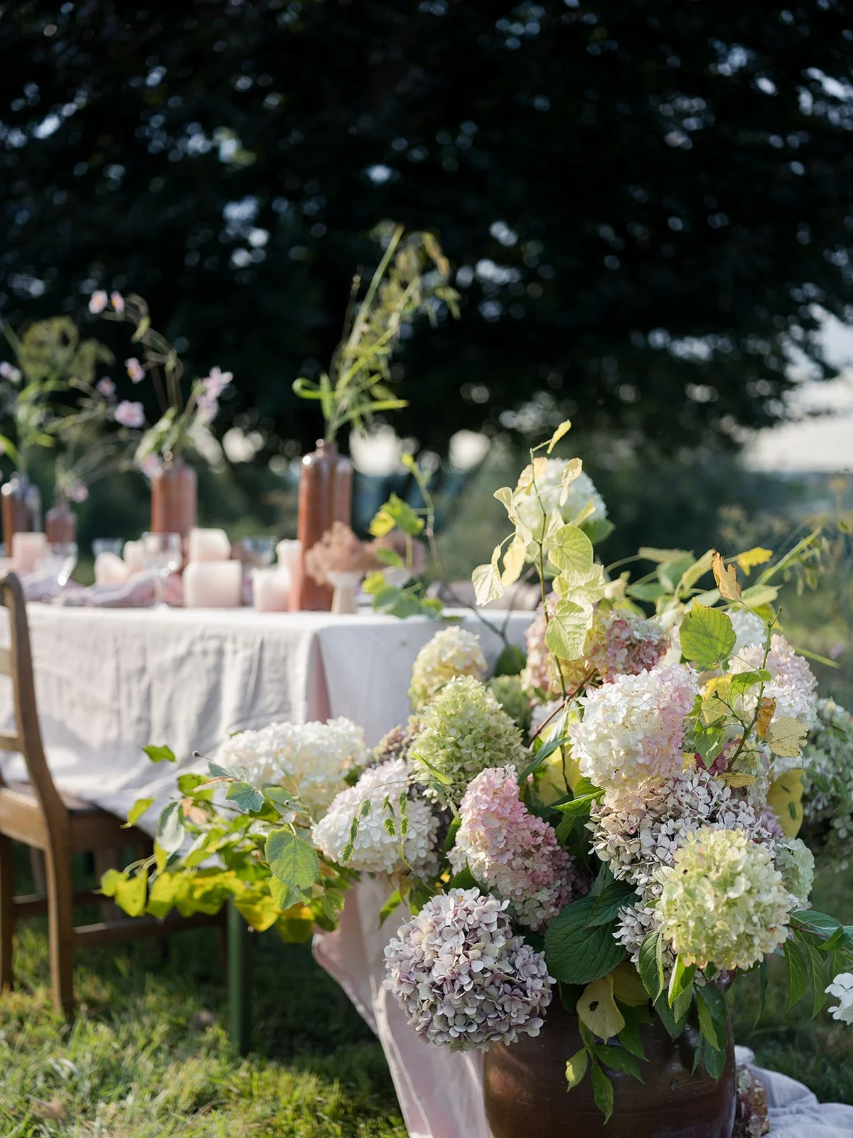 A large bouquet of white and pink hydrangeas and green leaves placed in front of a dining table decorated with candles and flowers on a grassy outdoor setting.