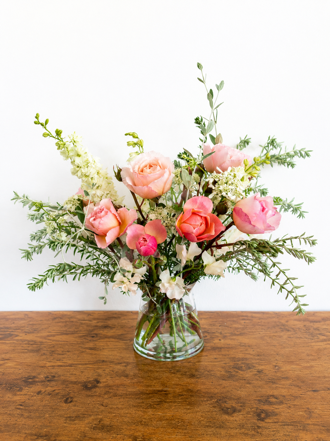 A glass vase with pink roses, white flowers, and green foliage on a wooden table against a white wall.