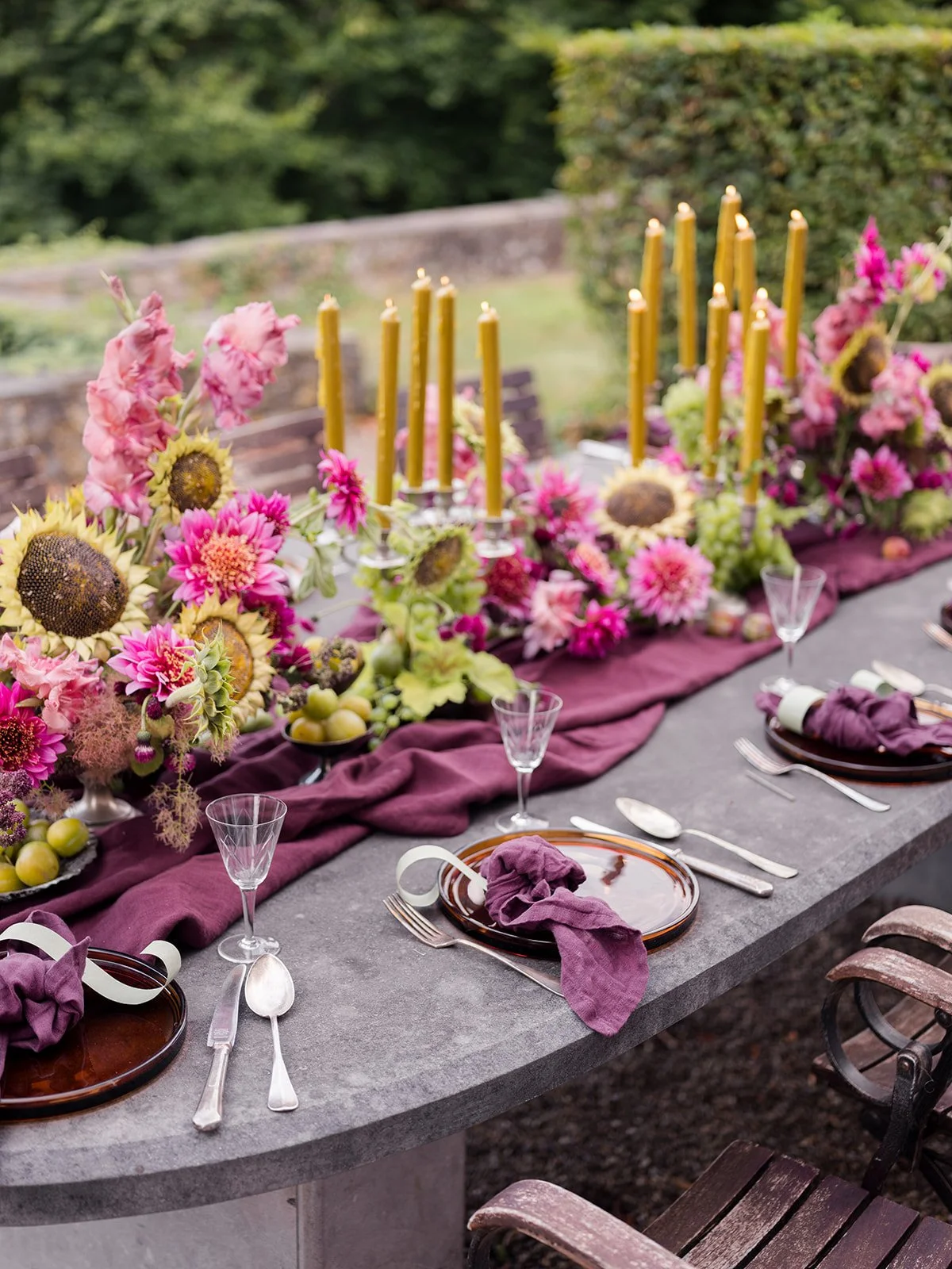 An outdoor dining table decorated with pink, yellow, and green floral arrangements, tall yellow candles, and purple tablecloths set with plates, glassware, and cloth napkins.