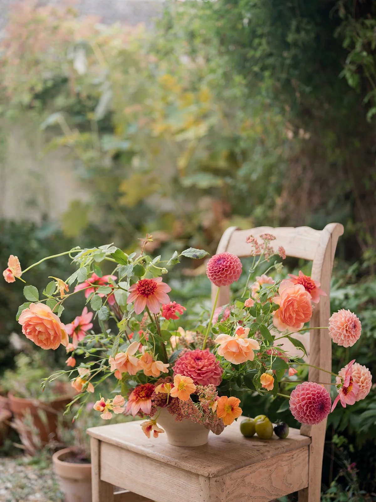 A bouquet of pink, orange, and yellow dahlias in a ceramic vase on a wooden chair.