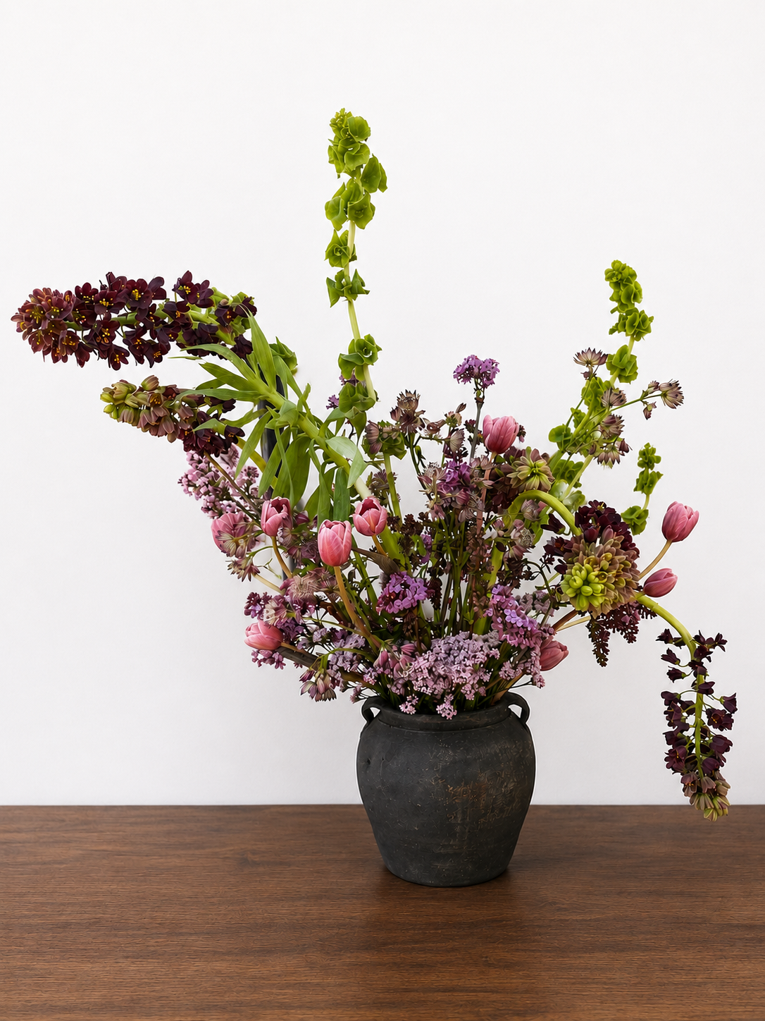 A Statement arrangement from Bloom House Collective in Marin County in a black vase with a variety of pink, purple, and green flowers arranged on a wooden table against a plain white wall.