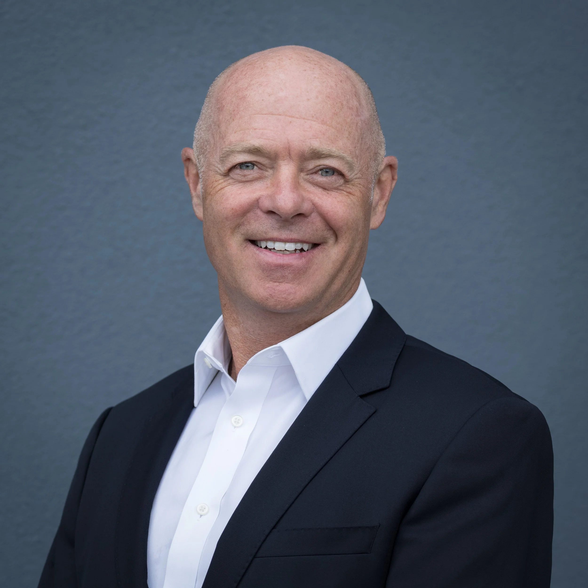 Close-up image of a smiling bald man wearing a tuxedo and a white shirt against a plain background.