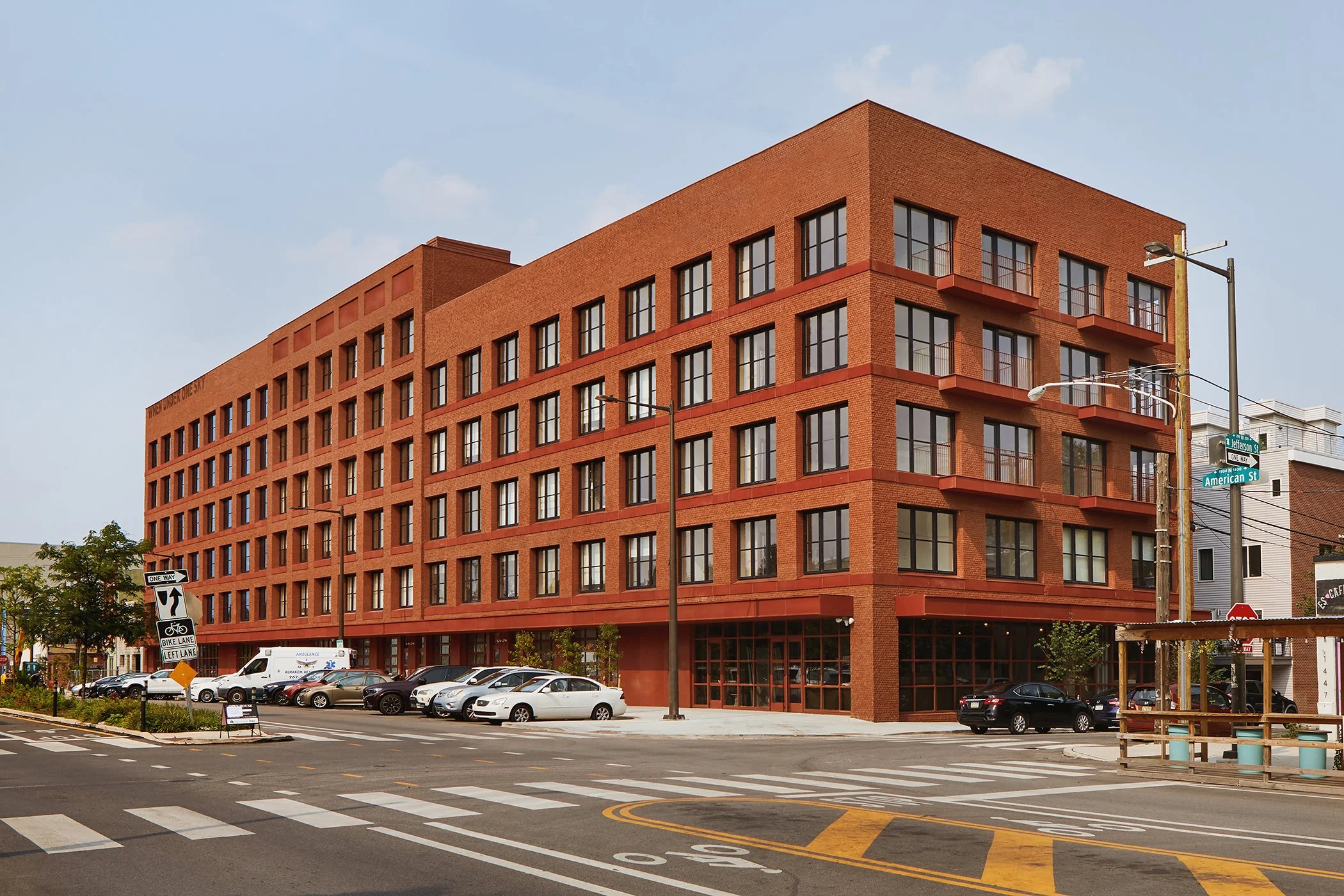Red brick multi-story building with large windows on a city street, with cars parked at the curb, street signs, and a crosswalk in the foreground.