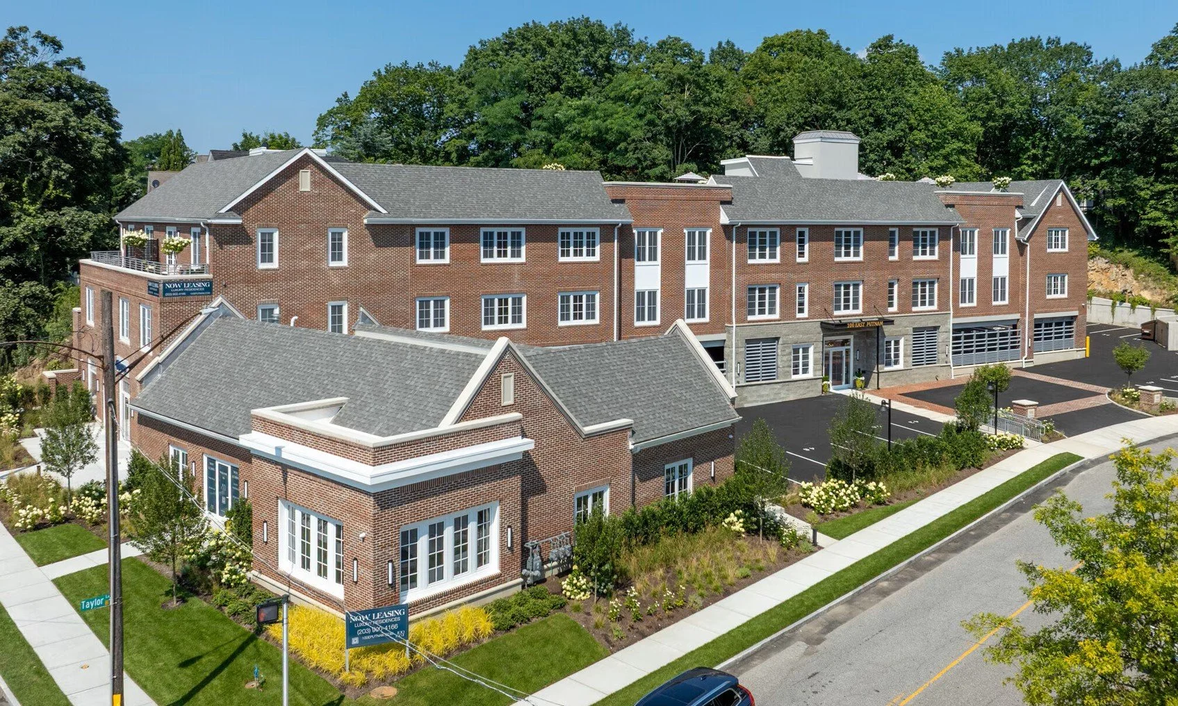A modern multi-story residential building complex with brick facades, multiple windows, and landscaped front yards, situated in a suburban neighborhood with trees and a clear blue sky.