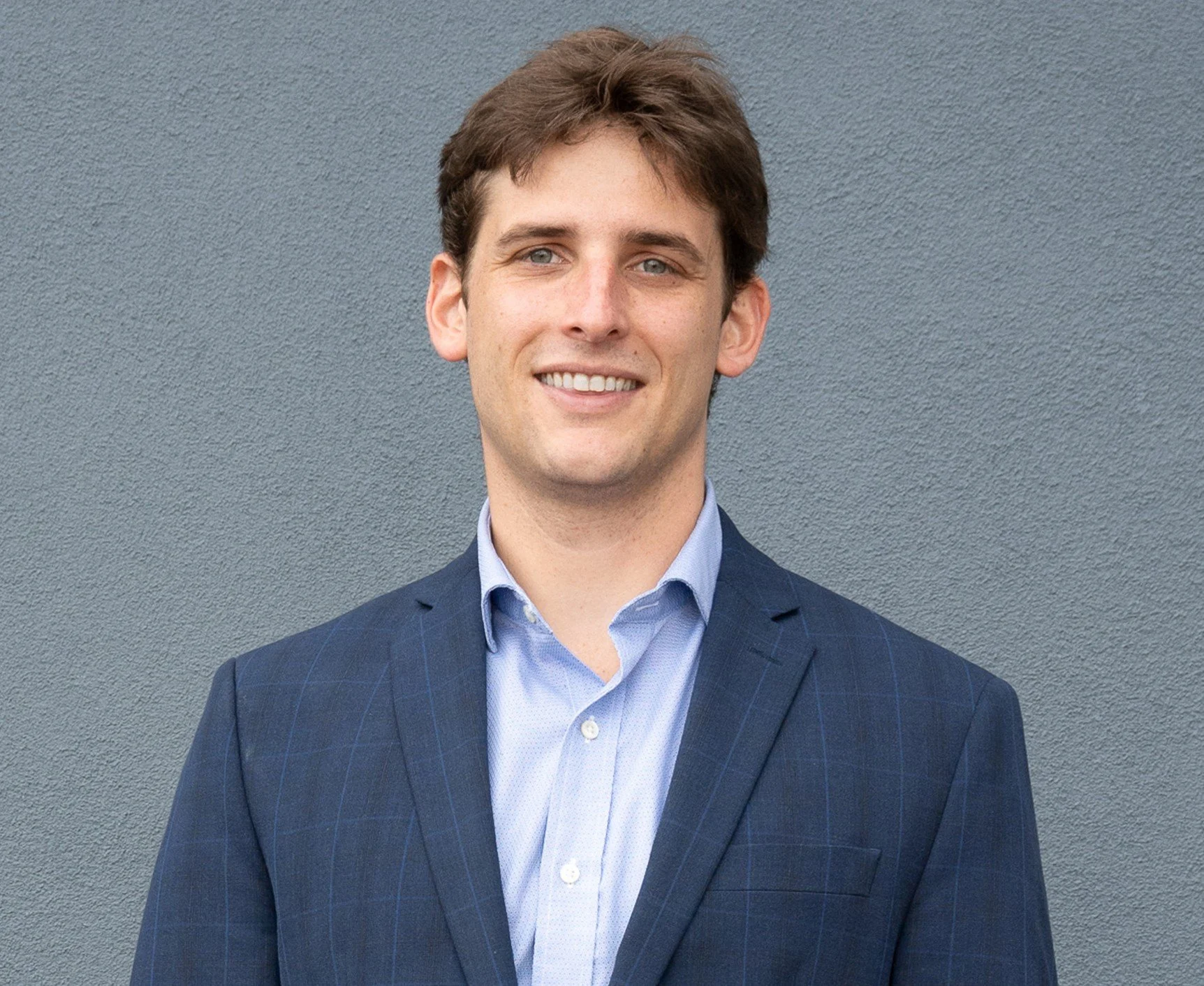 A young man with short brown hair, blue eyes, and fair skin, wearing a navy blue suit jacket and light blue button-up shirt, smiling in front of a plain gray wall.