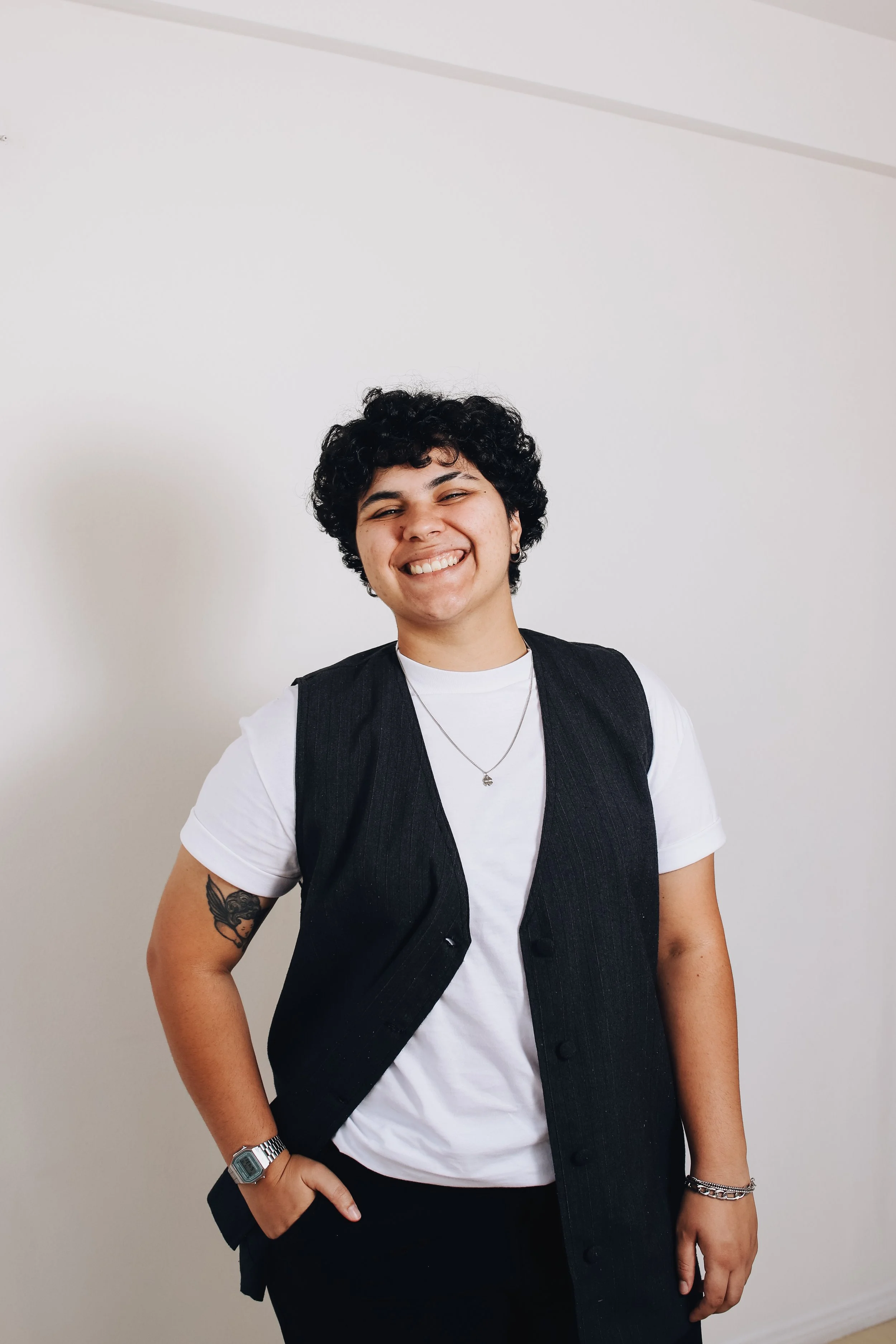 Jovem sorridente com cabelo cacheado escuro, vestindo uma camiseta branca, colete de risca de giz preto, usando relógio digital na esquerda e colar, posando com a mão na cintura, against uma parede branca.