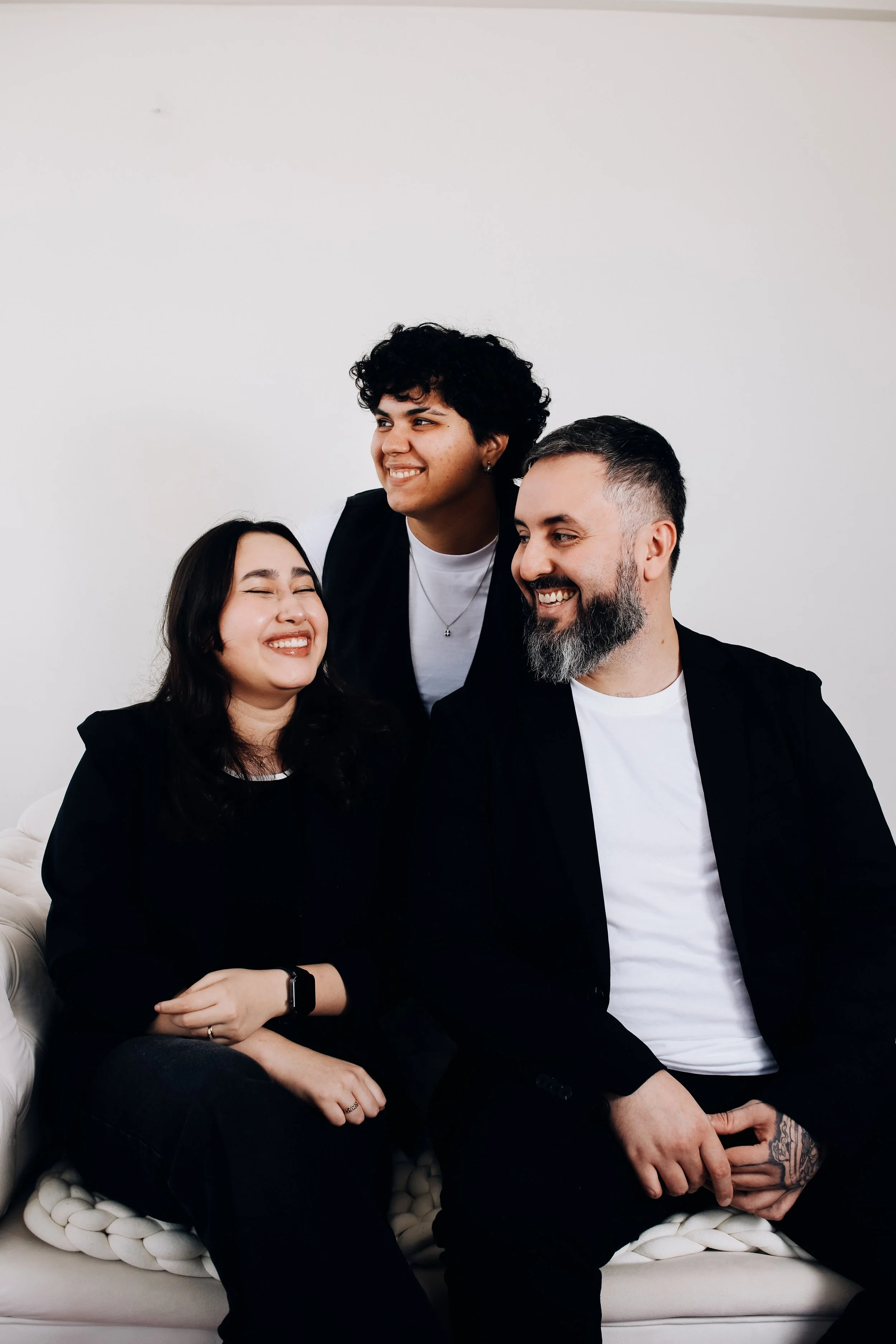 Três pessoas sorrindo sentadas e uma pessoa de pé, todos com roupas casuais, em um ambiente com parede branca ao fundo.