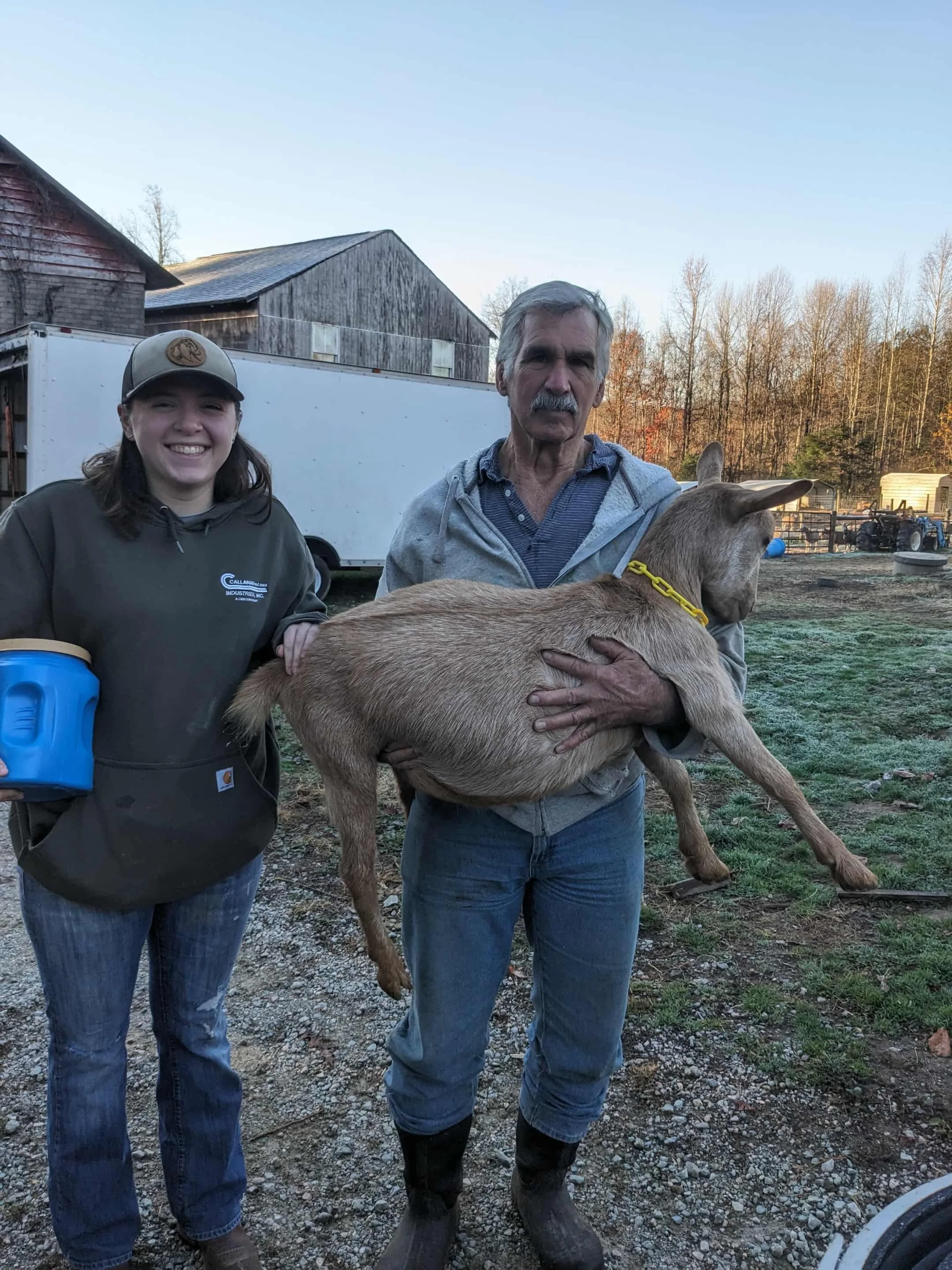 A man holding a baby goat and a young woman standing nearby, on a farm with barn and trees in the background.