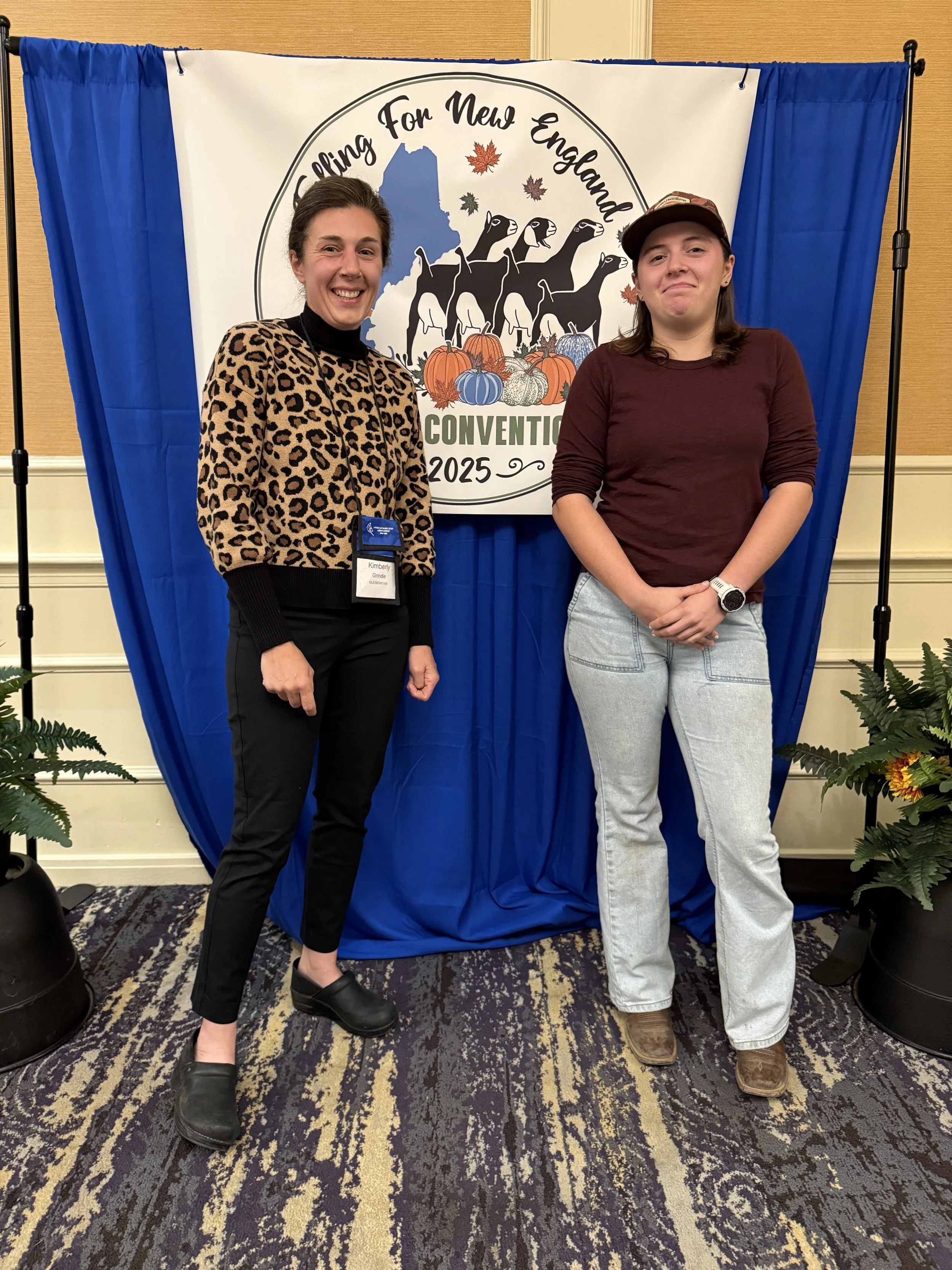Two women standing in front of a conference backdrop with a logo depicting dogs, pumpkins, and fall leaves, at a convention in 2025.