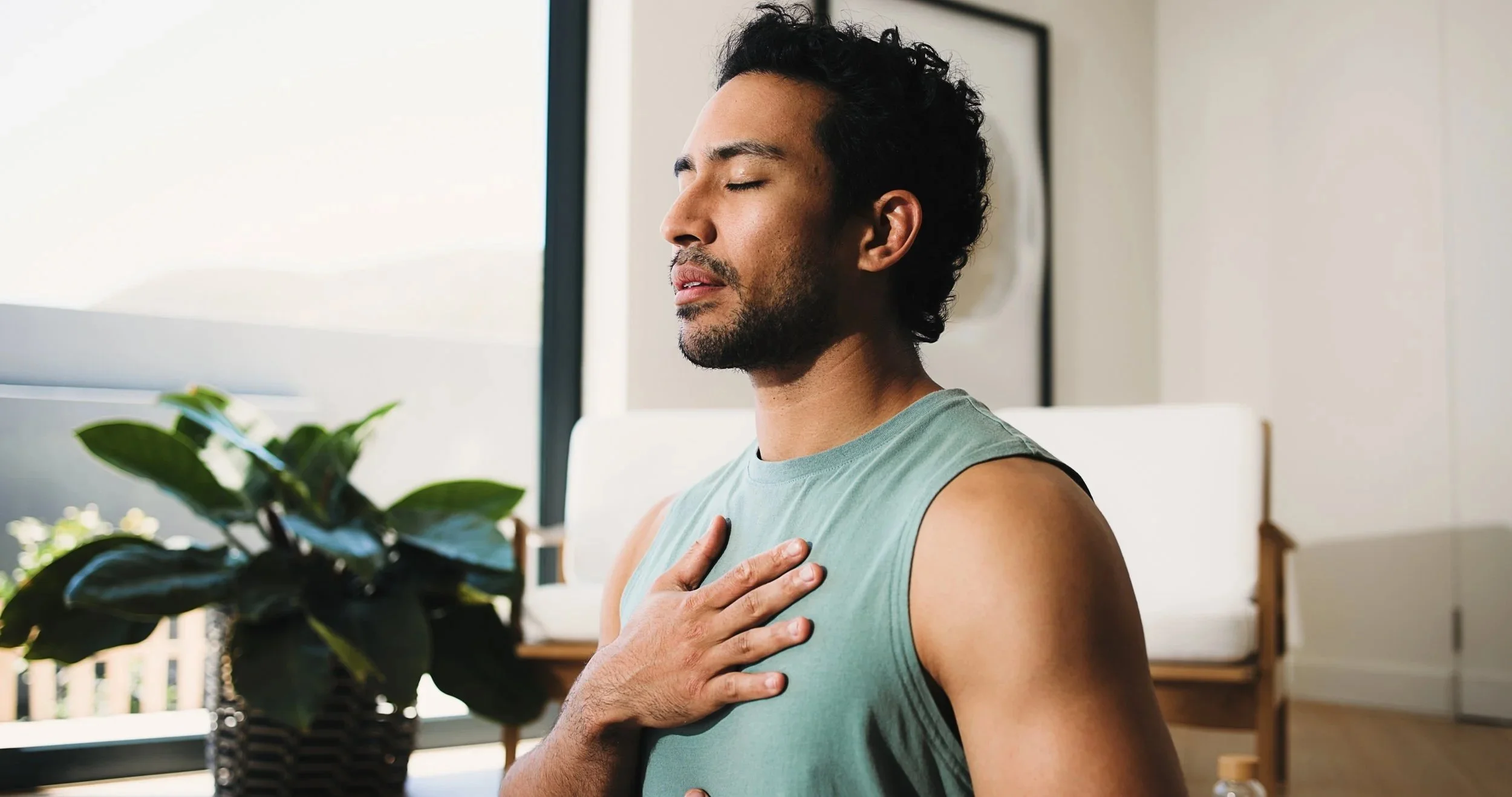 A man with closed eyes and a hand on his chest, practicing meditation or mindfulness in a bright room with a large window and a plant nearby.
