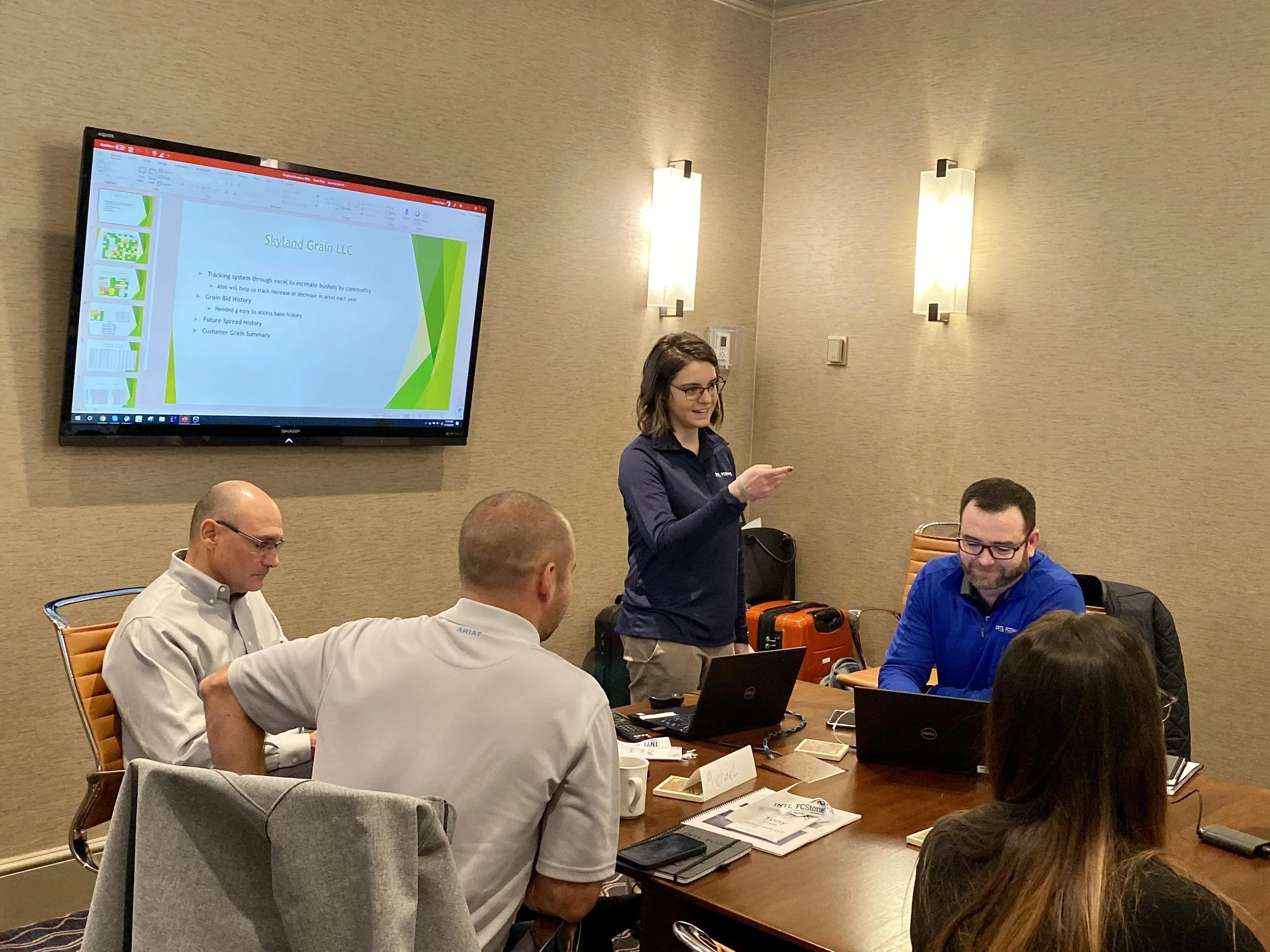 A woman standing at a conference table giving a presentation to four seated people. The woman is looking at her phone, and a PowerPoint slide is projected on a large TV screen behind her.