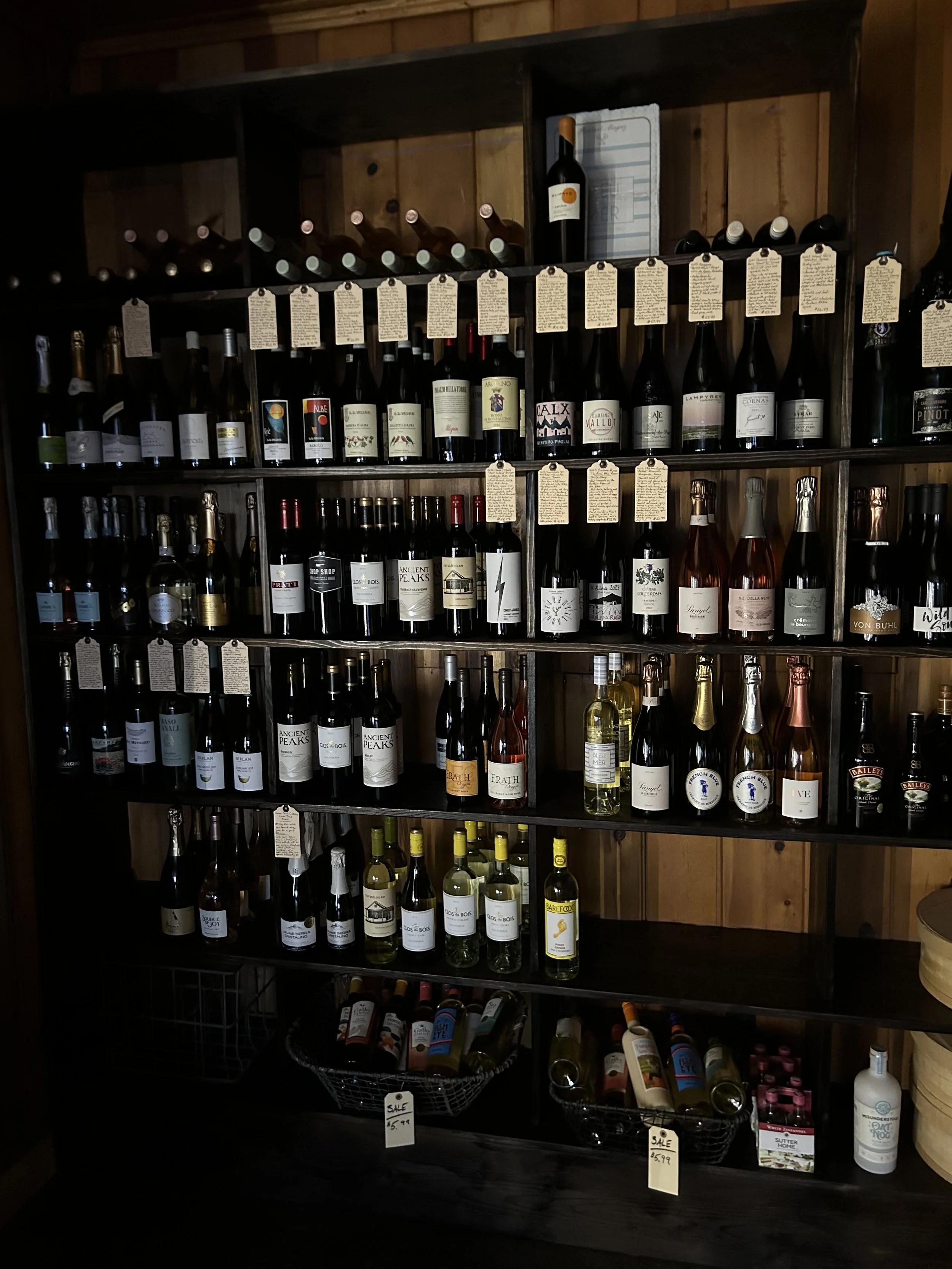 A wooden wine rack filled with various bottles of wine, with handwritten tags hanging from some bottles, located in a cozy, wood-paneled environment.