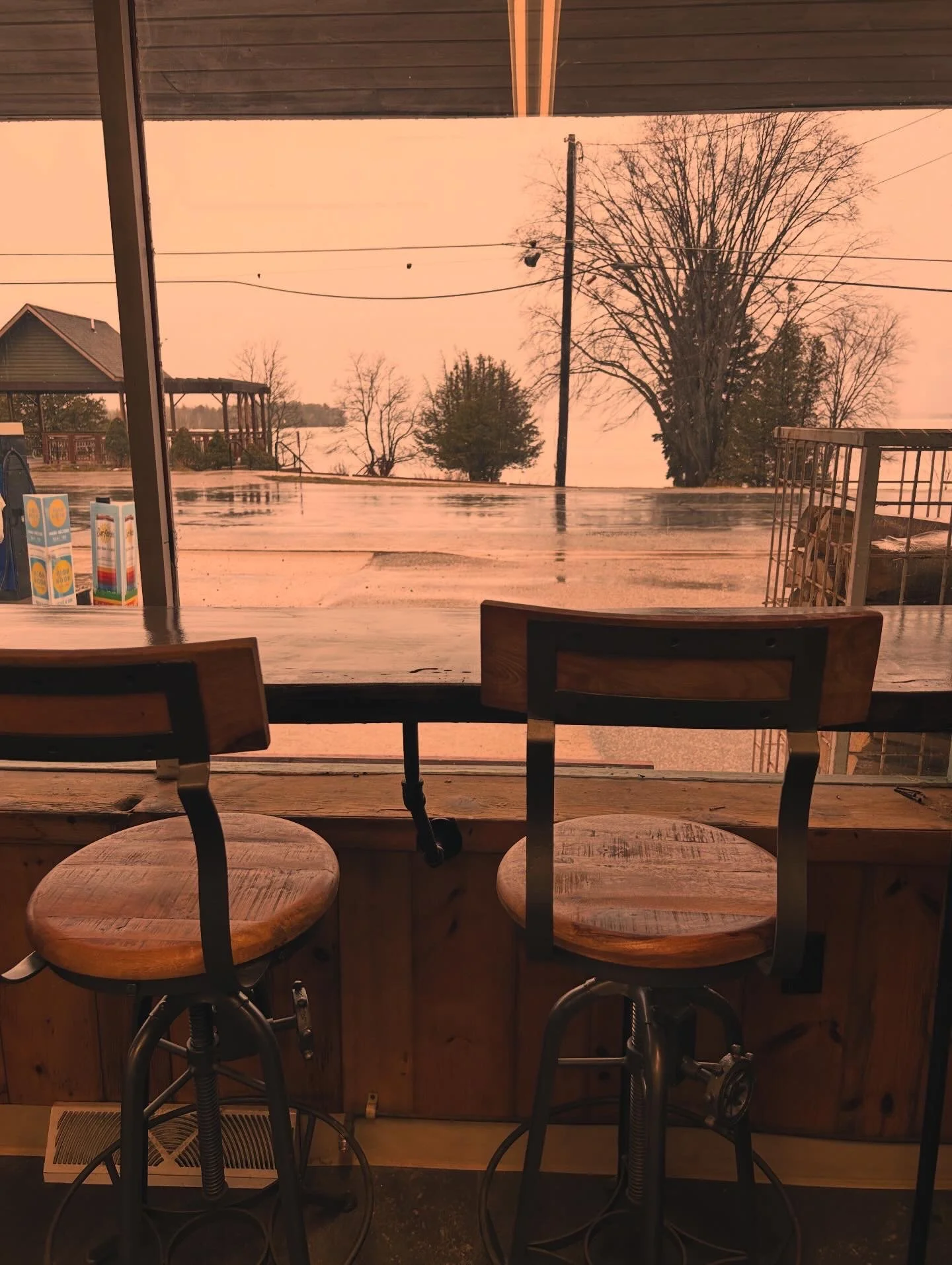 View from inside a restaurant or cafe window, showing two wooden bar stools at a counter, with a wet outdoor scene including trees, a utility pole, and power lines on a rainy day in the distance.