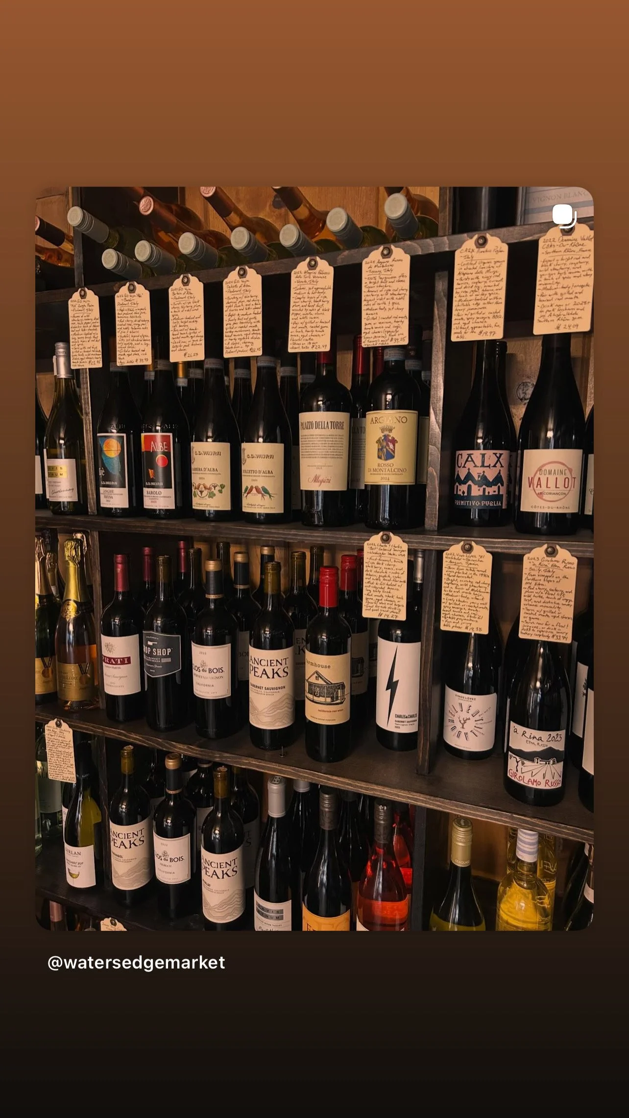 Shelf of assorted wine bottles with handwritten tags, in a wine shop or bar setting.