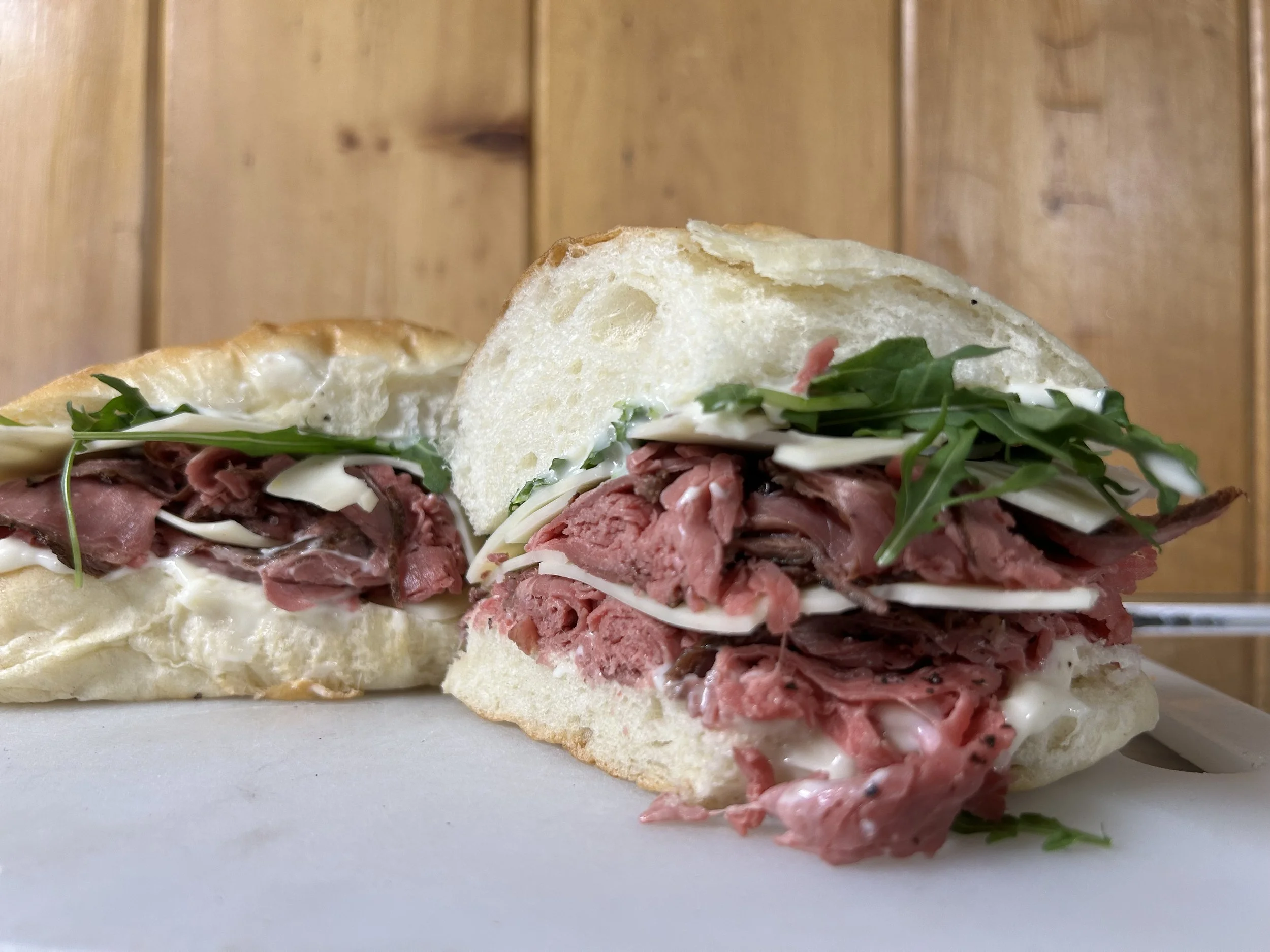A sliced roast beef sandwich with melted cheese, arugula, and sliced white onions on a crusty white bread roll.