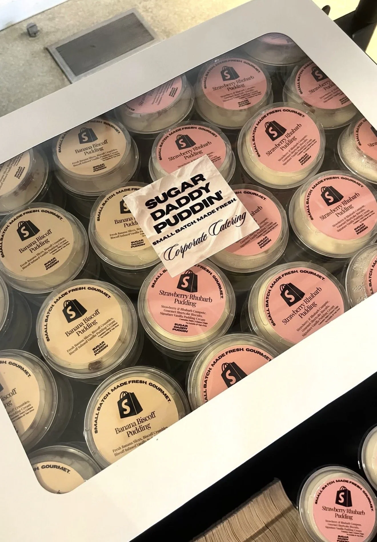 A white box containing multiple small containers of pudding, with flavors including banana biscuff and strawberry rhubarb, arranged in rows with a label from Sugar Daddy Puddin' on top.