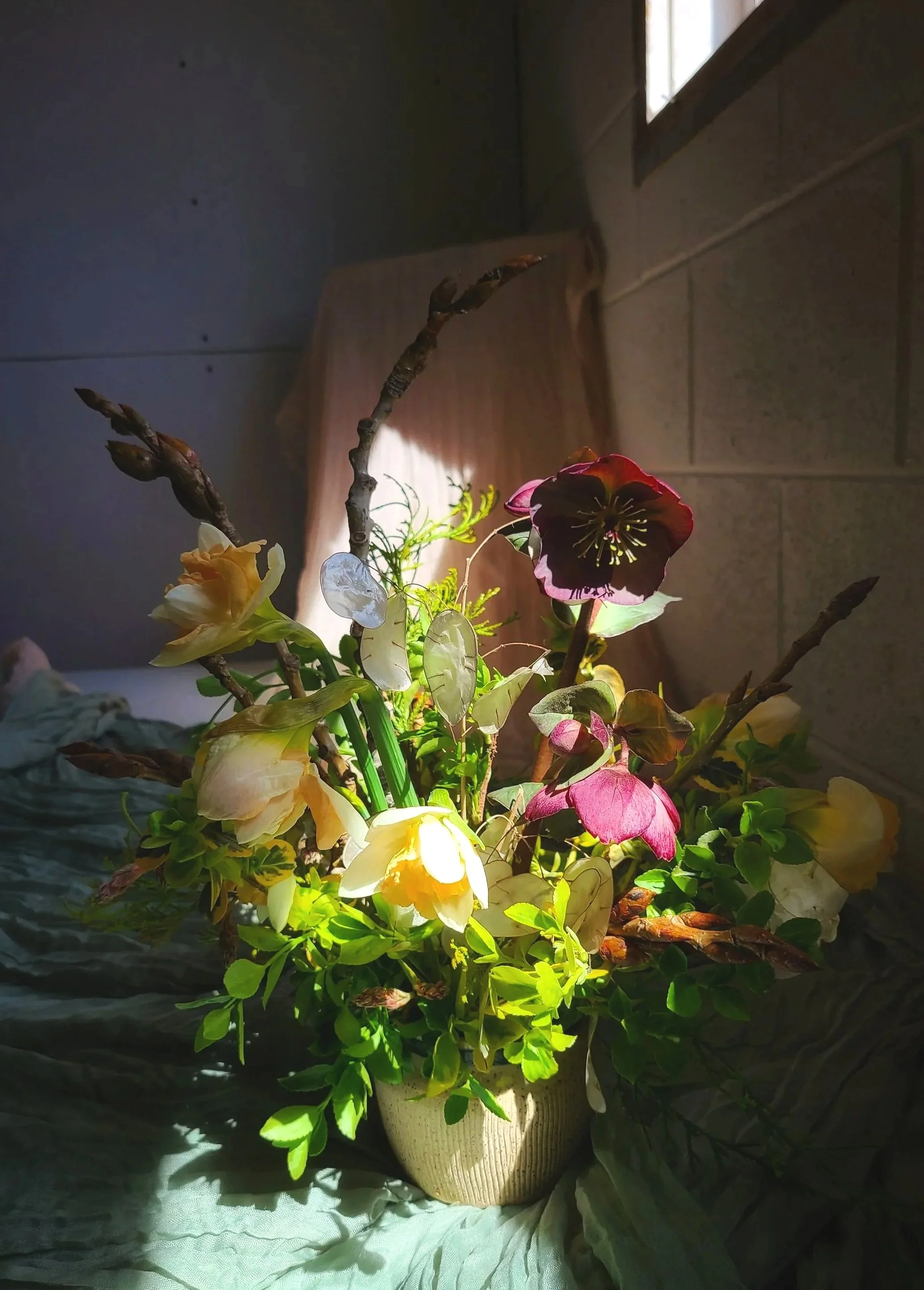 A floral arrangement in a white ceramic vase with various flowers and green foliage, illuminated by a light source from above.