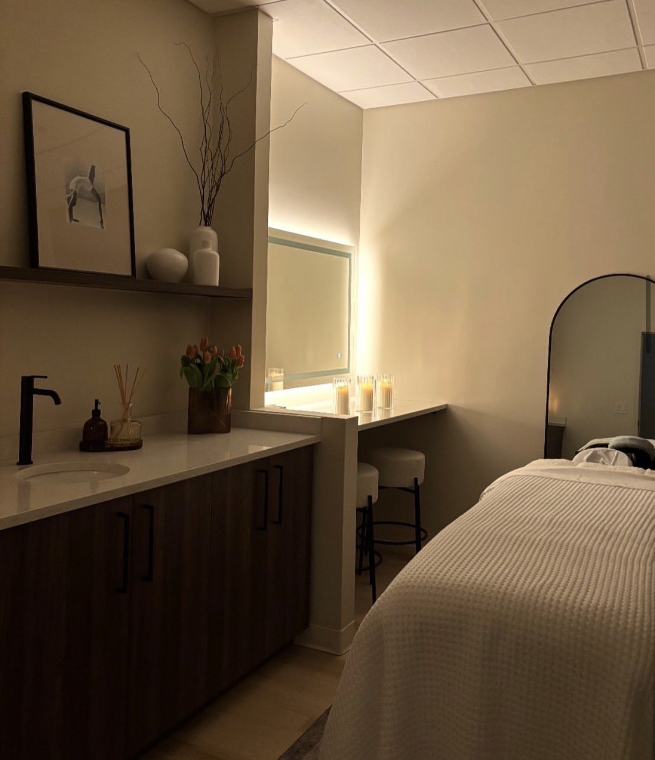 A cozy hotel room with a bed, a small kitchen area with a sink, decorative items including a framed picture of a deer, white vases, a small potted plant, and candles on a ledge near a window with soft lighting.