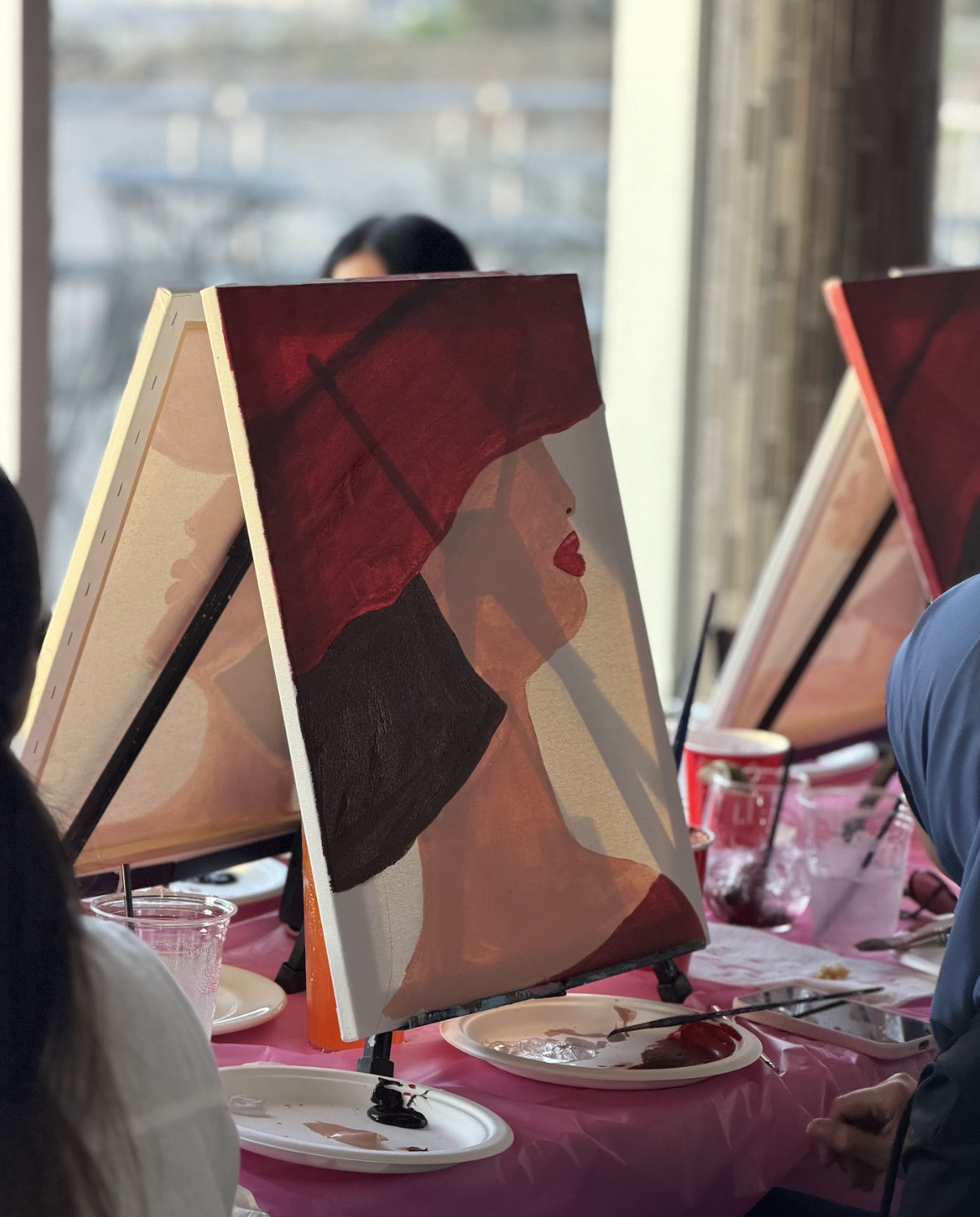 Painting of a woman with red lips, wearing a red umbrella hat, and black clothing, on an easel at an art event.