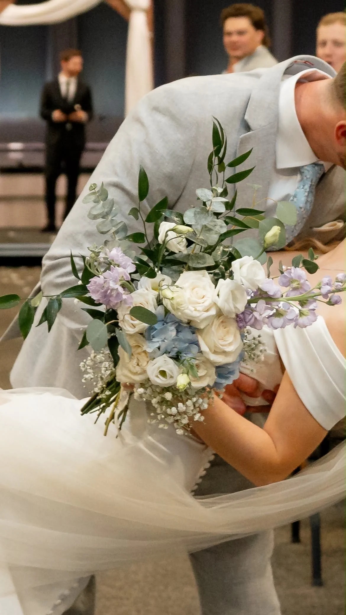 A person holding a bouquet of white, purple, and light blue flowers with green leaves, possibly at a wedding or special event.