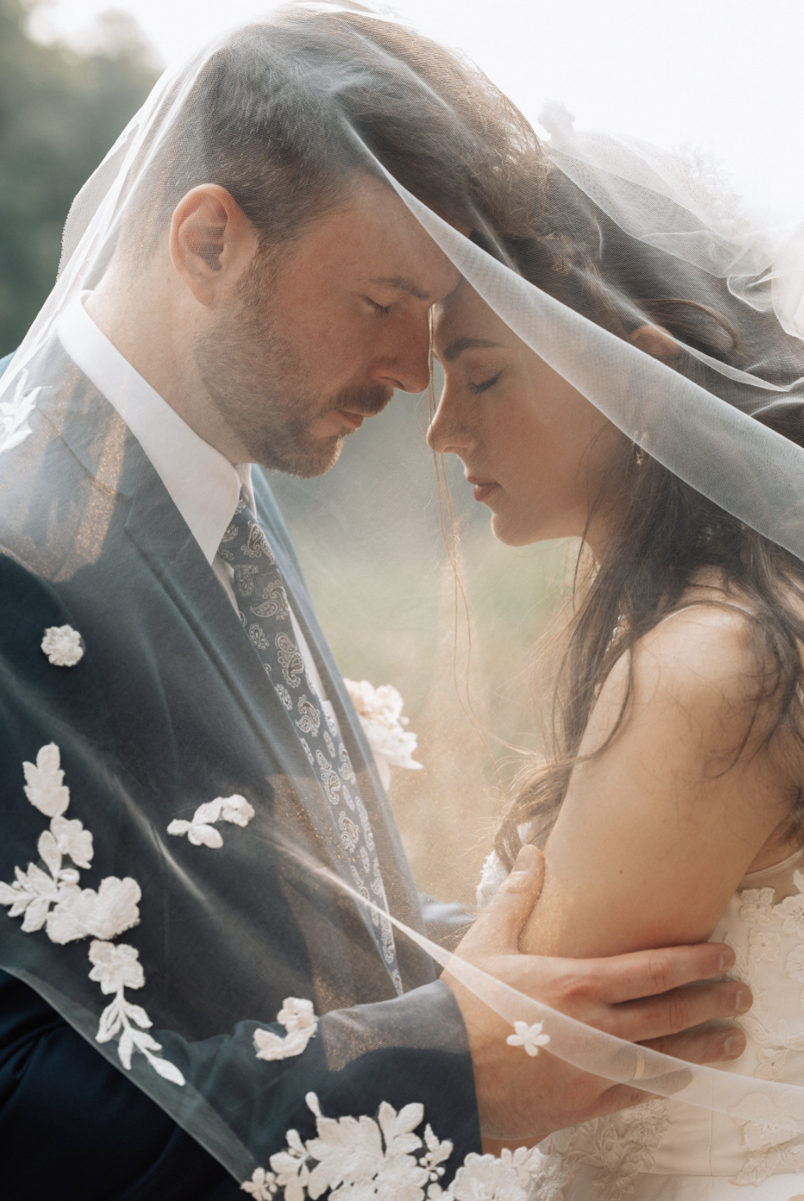 A bride and groom with foreheads touching, embracing under a wedding veil outdoors during sunset.
