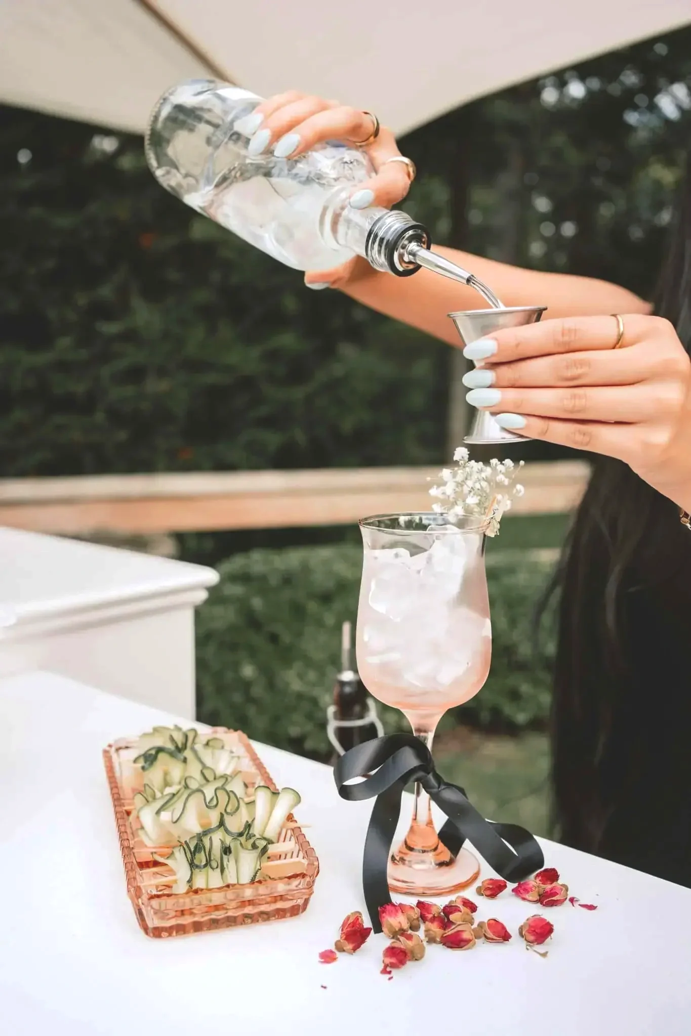 A person pours water into a glass with flowers inside at an outdoor setting, with assorted sliced cucumbers and dried rose petals on a white tablecloth.