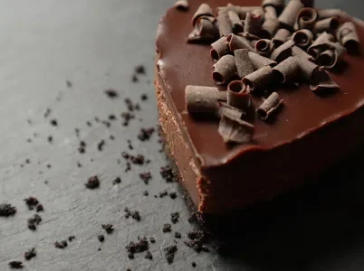 A close-up of a heart-shaped chocolate cake topped with chocolate curls, with scattered crumbs on a gray surface.