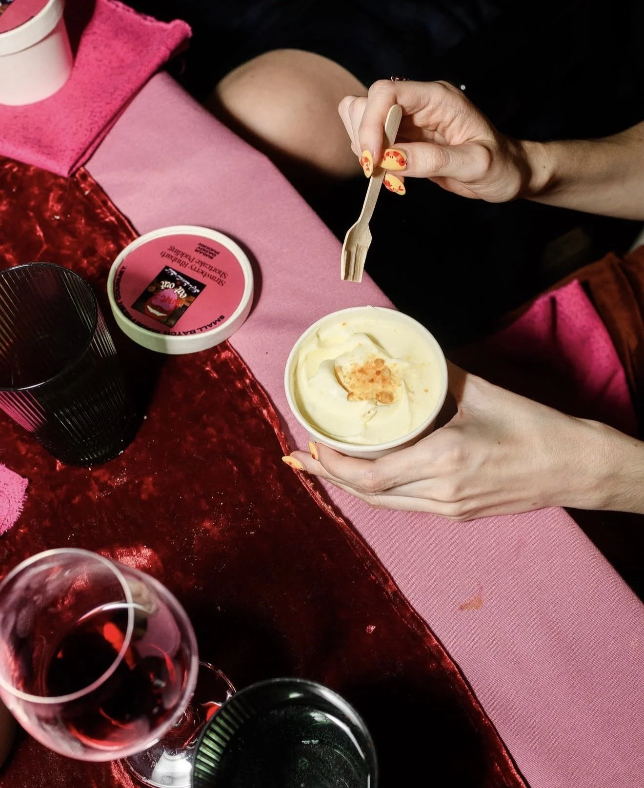 A person with painted nails holding a small wooden fork and eating ice cream in a cup, at a table with wine glasses, a pink and red cloth, and a container of strawberry ice cream.