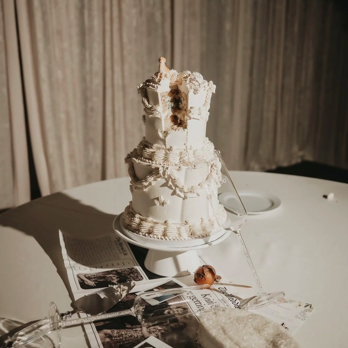 A partially eaten white wedding cake with three tiers on a white cake stand, situated on a table covered with a white tablecloth. The table also has scattered photos, a magazine, and a cake knife.
