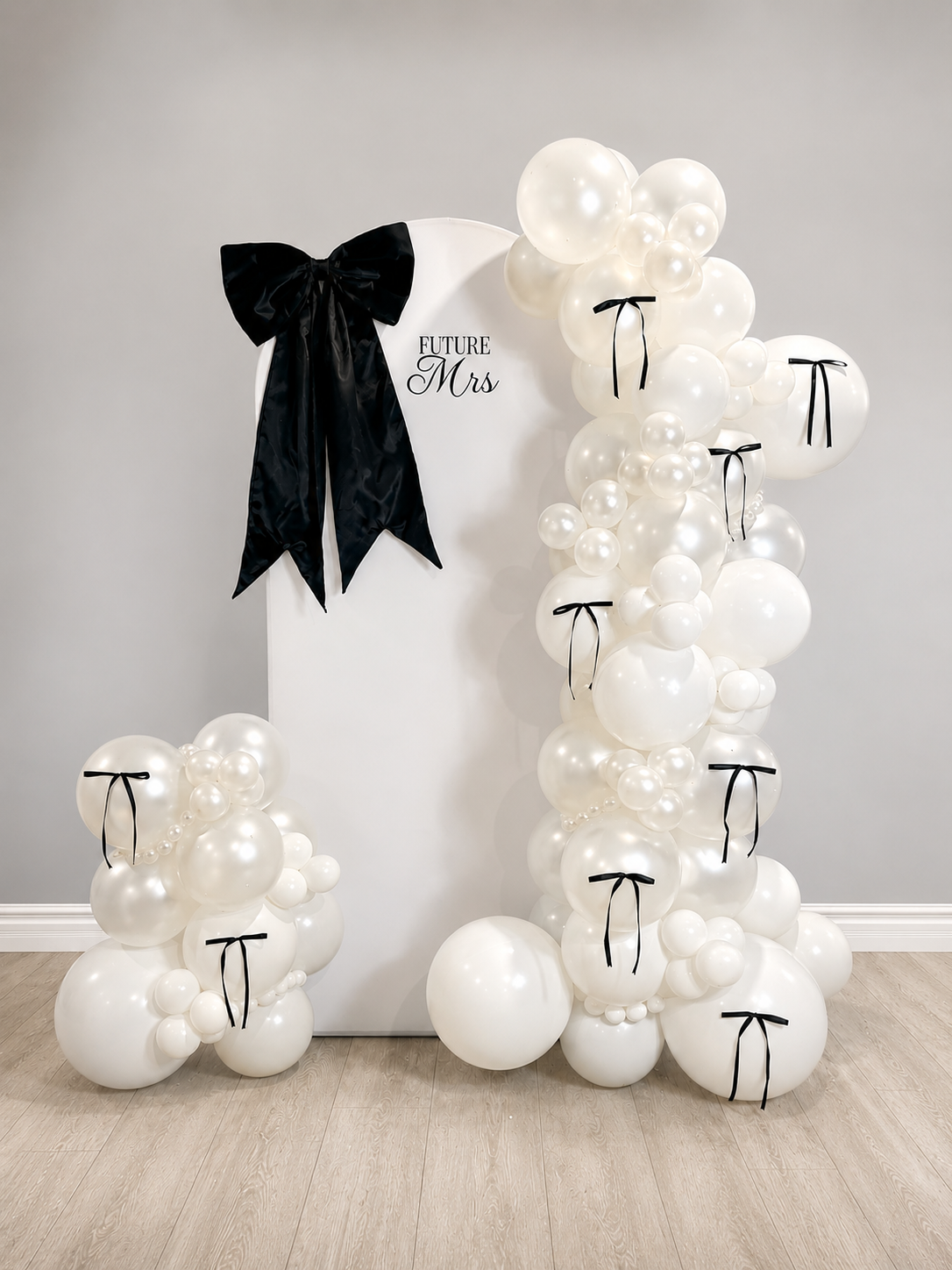 Decorated backdrop with a white panel, black bow, and the words 'Future Mrs.' on it, surrounded by an arch of white balloons with black ribbons on some of them.