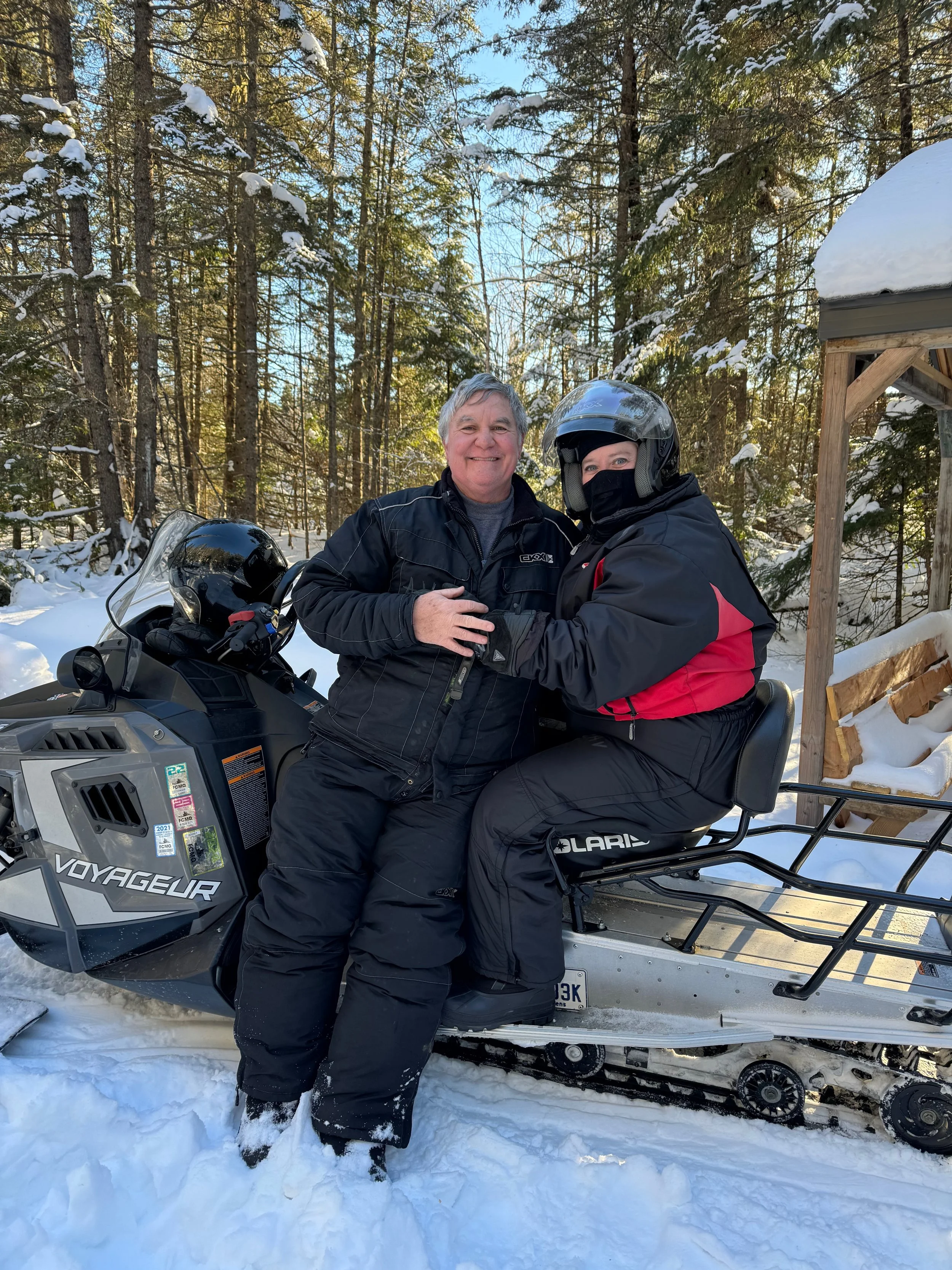Two people wearing winter gear, sitting on a snowmobile in a snowy forest with evergreen trees and a wooden shelter visible in the background.