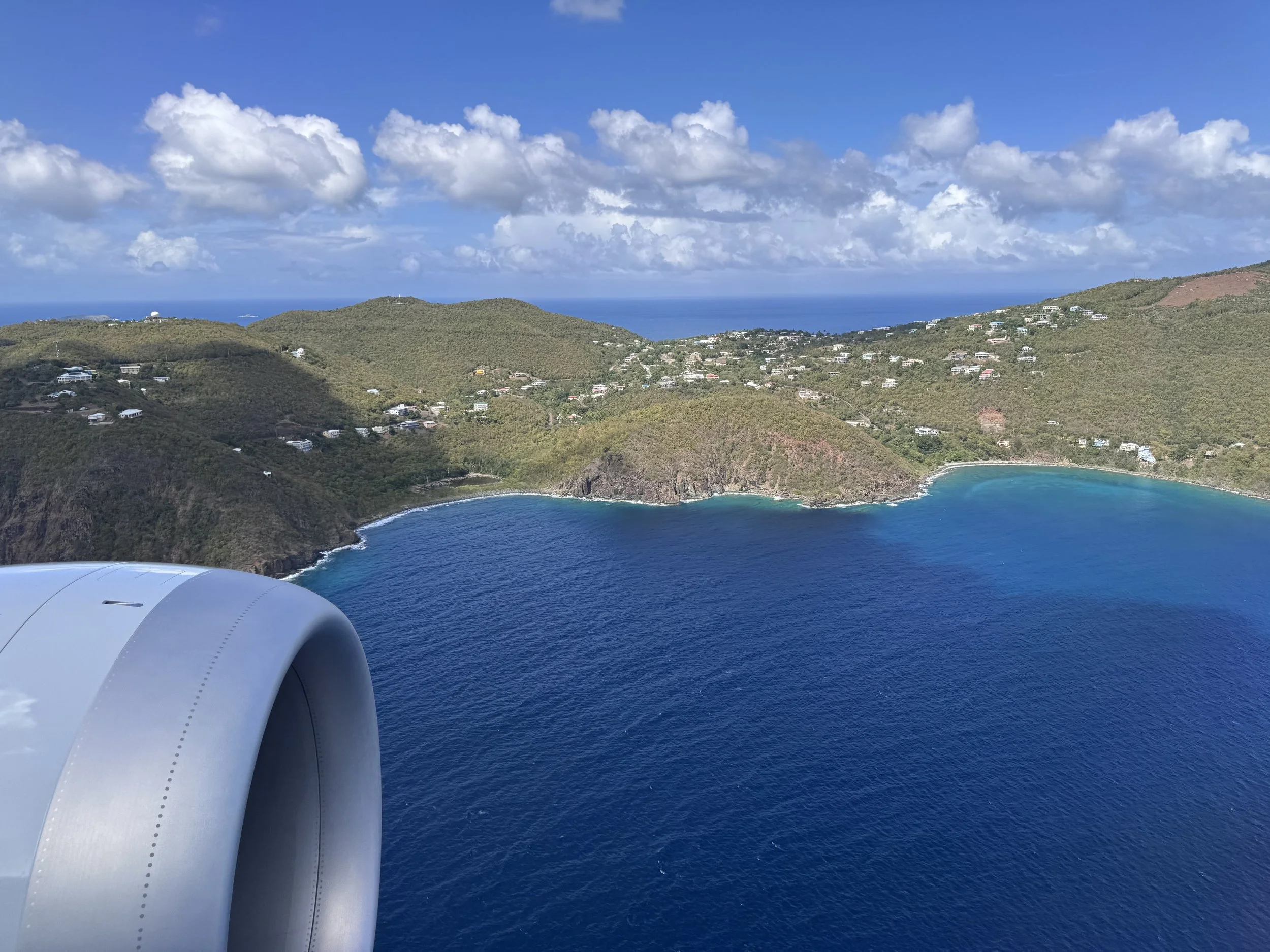 View from an airplane window showing a blue ocean coastline, lush green hills with scattered houses, and a partly cloudy sky.