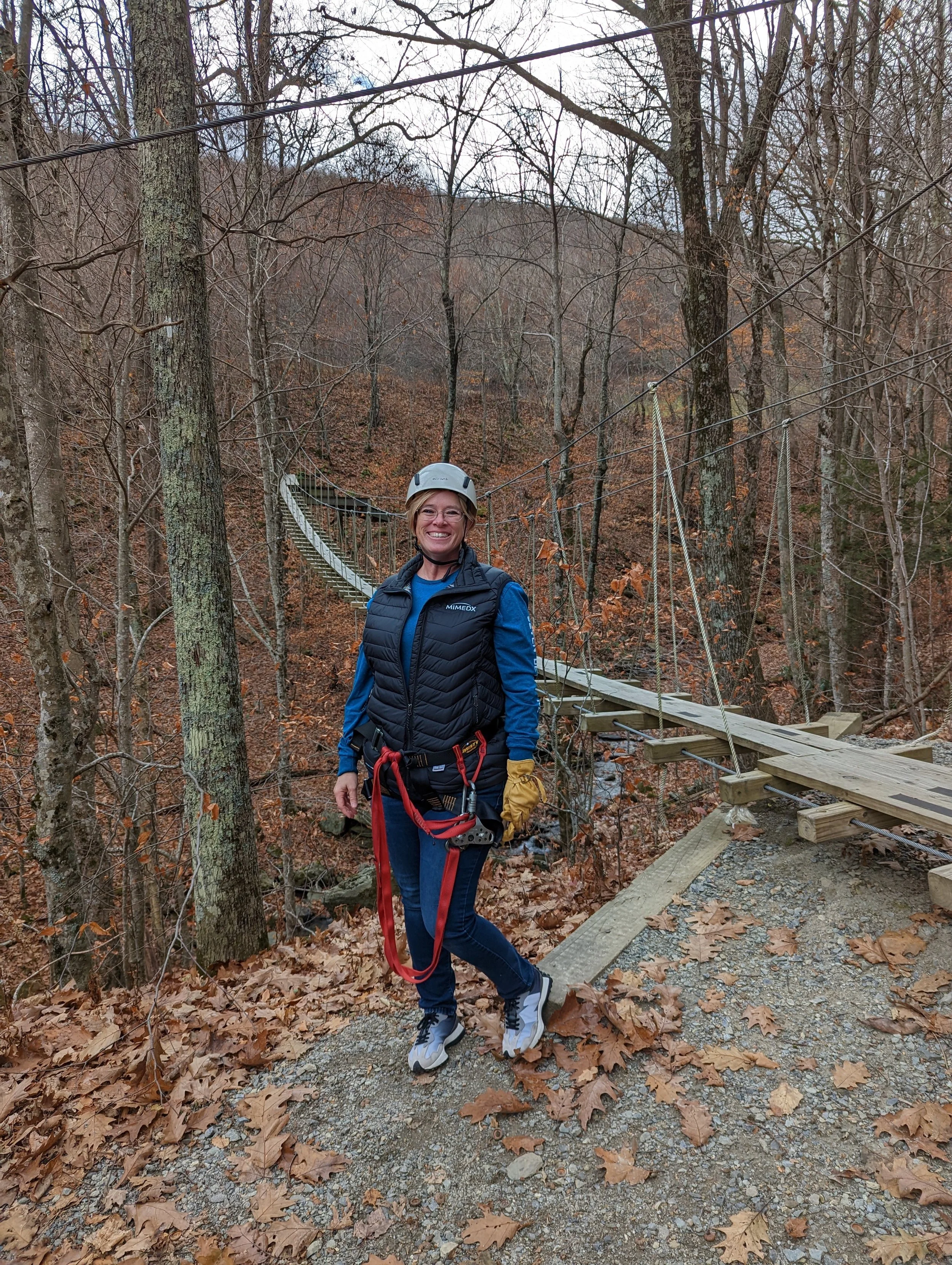Woman wearing safety gear standing on a trail near a suspension bridge in a wooded area with fallen leaves.