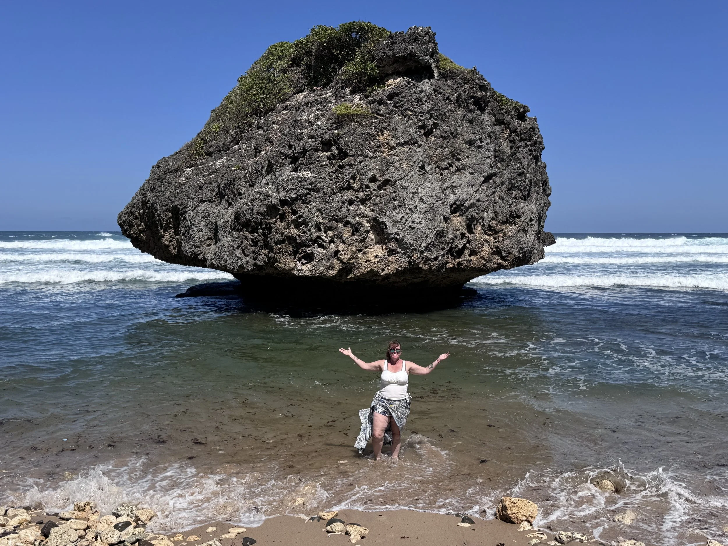 A woman standing in the ocean water with arms raised in front of a large rock formation on the beach. The sky is clear and blue with waves in the background.
