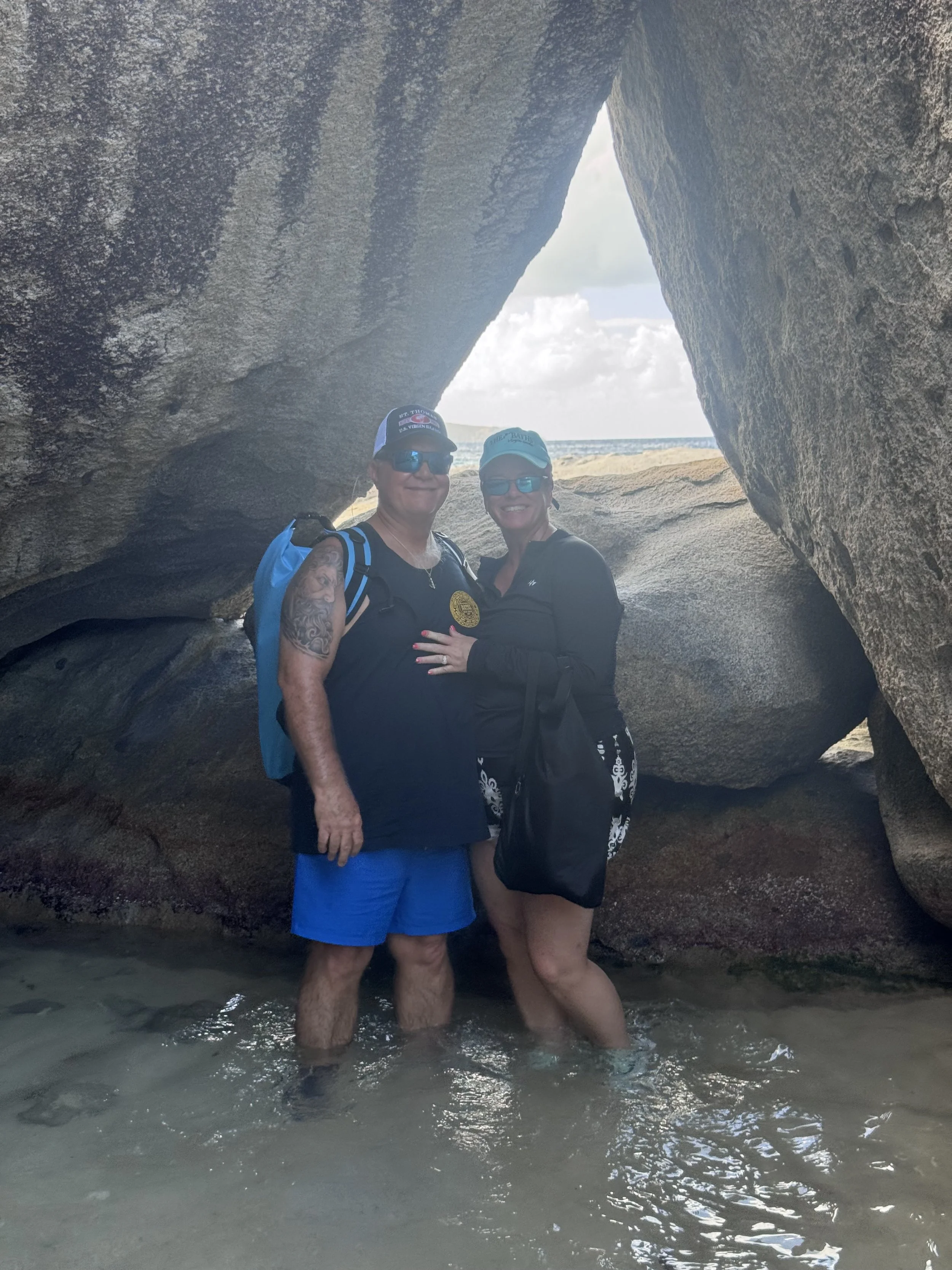 A man and woman standing in shallow ocean water beneath large rocks on a beach, smiling and posing for a photo.