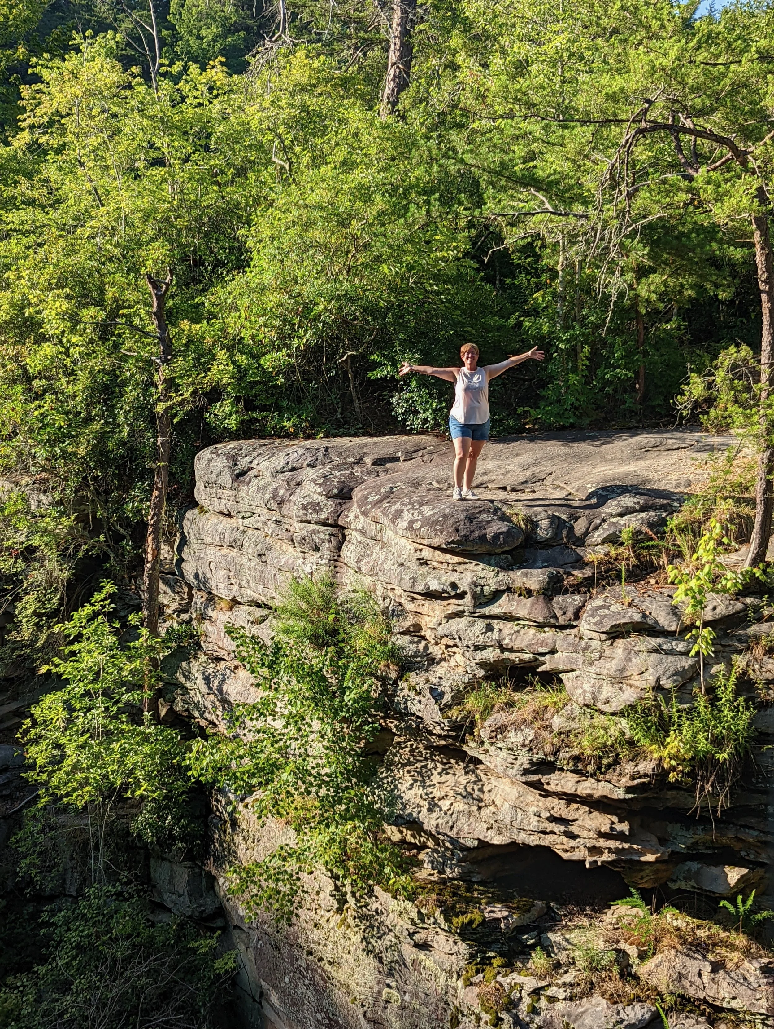 A woman stands with arms outstretched on a large rock ledge in a wooded area.