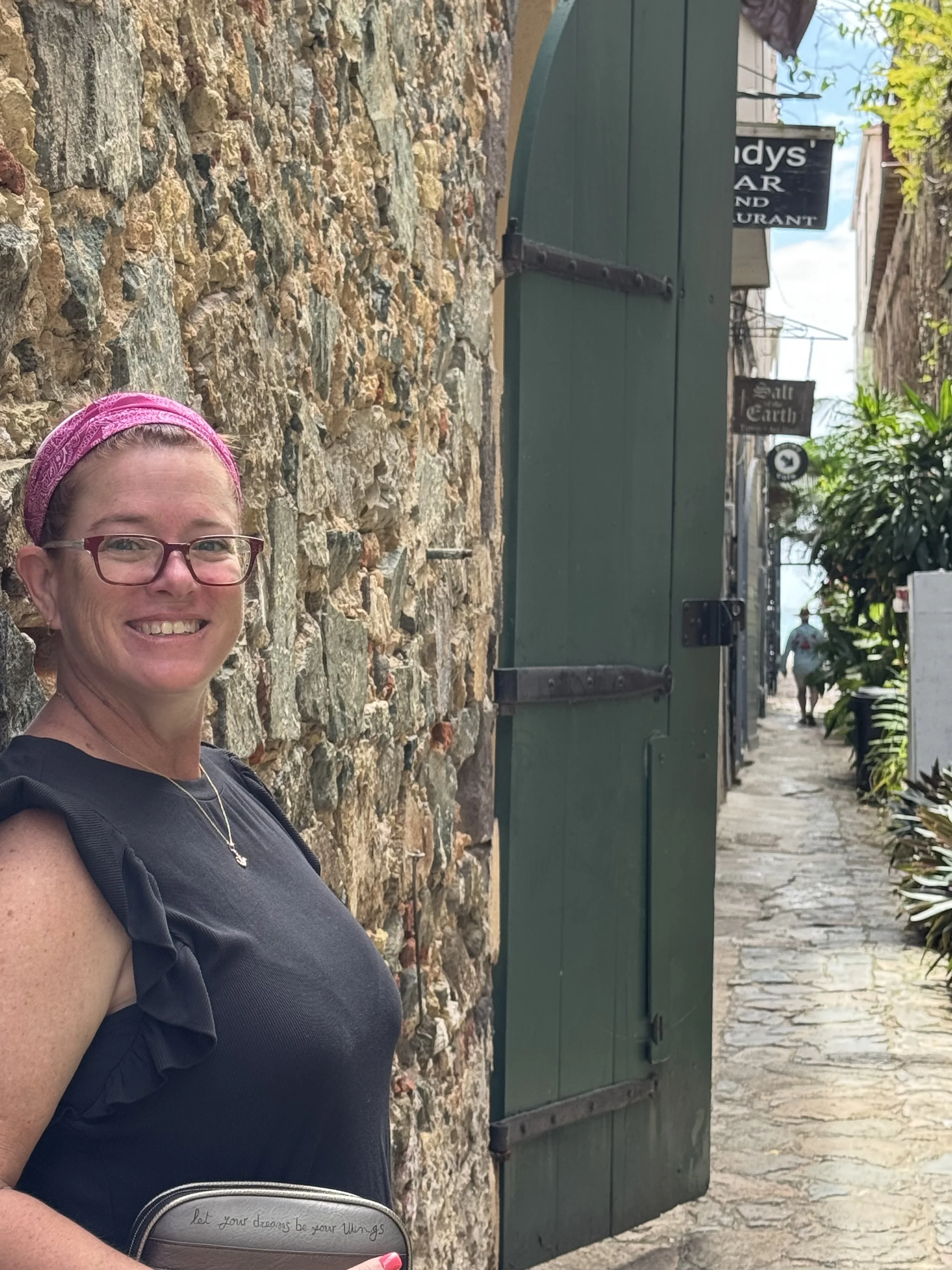 A woman with glasses and a pink headband smiling, standing next to a stone wall and green door, on a street with signs and trees.
