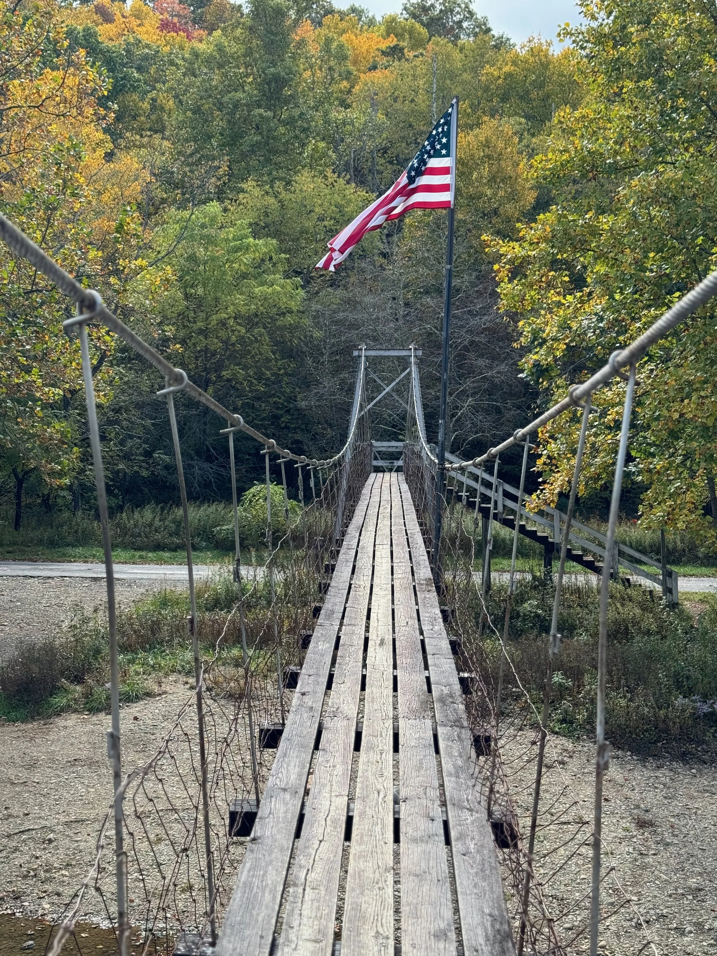 A suspension bridge made of wooden planks with metal railings on each side, spanning over a small creek with trees in the background. An American flag is flying on a pole above the bridge, amid fall foliage.
