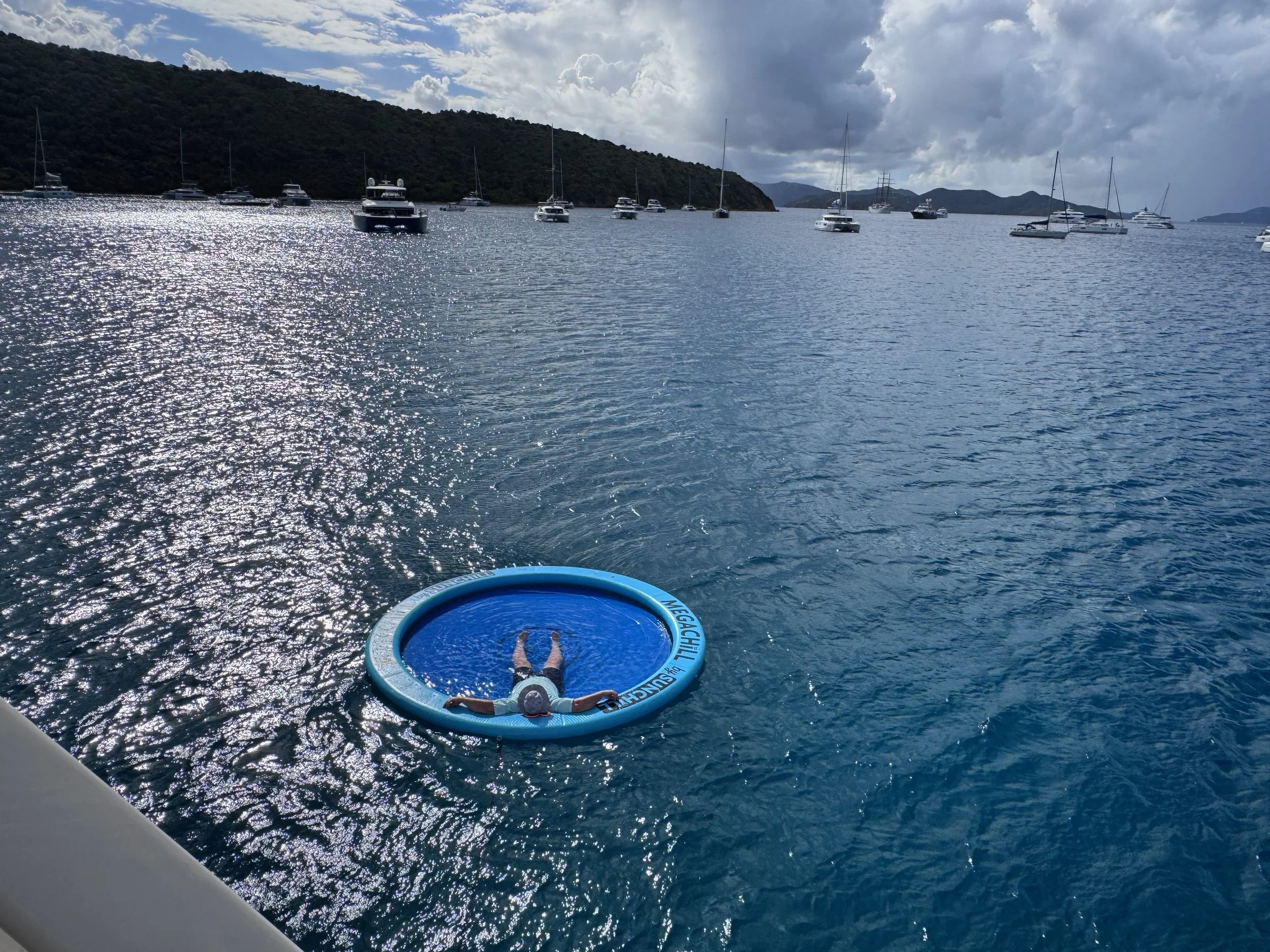 A person lying on a blue inflatable ring floating on a body of water, with a few smaller boats and sailboats anchored nearby, surrounded by hilly islands in the distance under a partly cloudy sky.