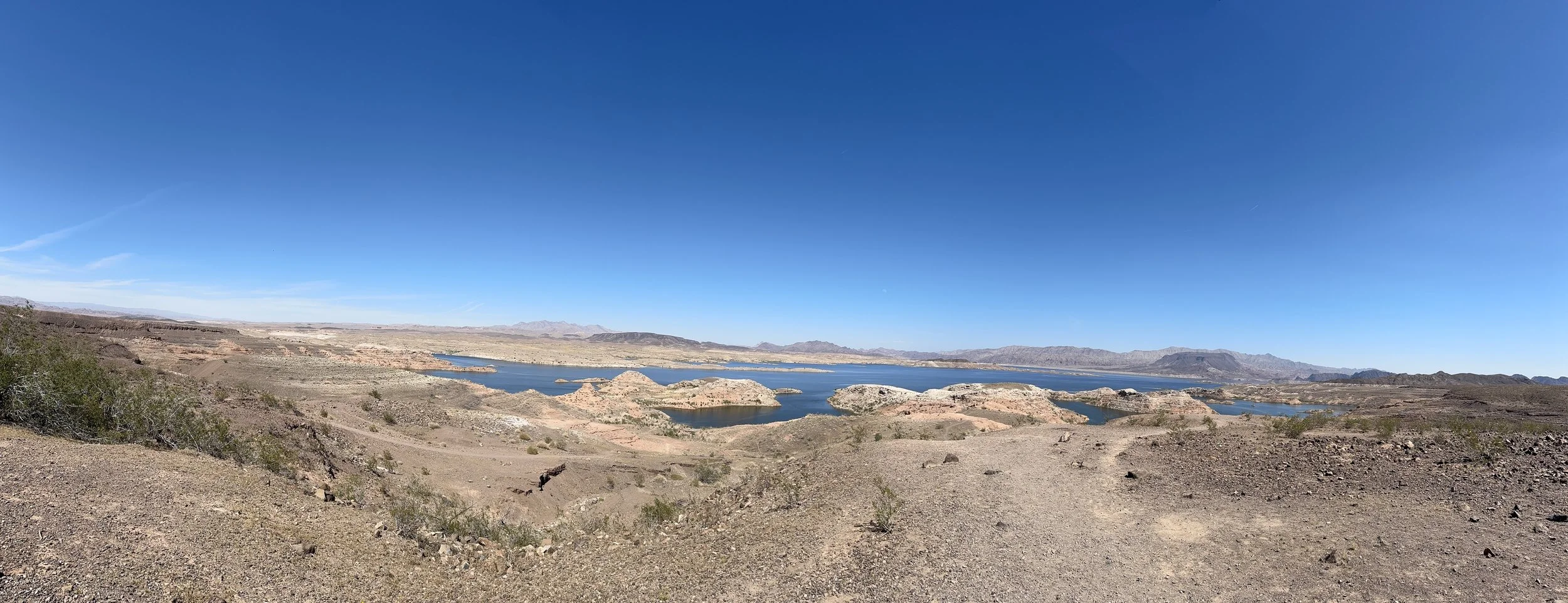 A panoramic view of a desert landscape with a large body of water, rugged terrain, and distant mountains under a clear blue sky.
