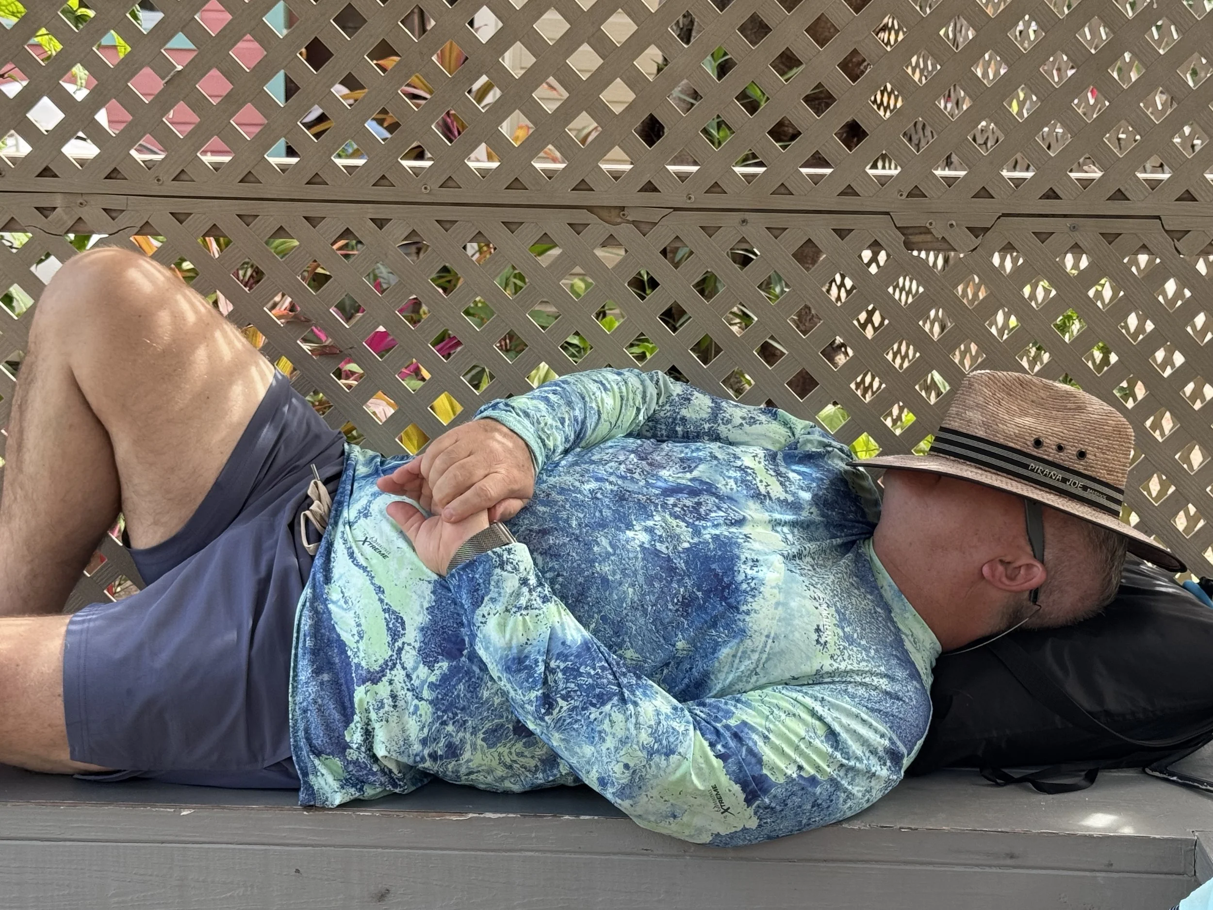 A person dressed in a colorful, patterned shirt and dark shorts, wearing a straw hat, is lying or reclining outdoors on a wooden surface. Part of the person's face and head are visible, and a few green plants can be seen behind a decorative lattice fence.