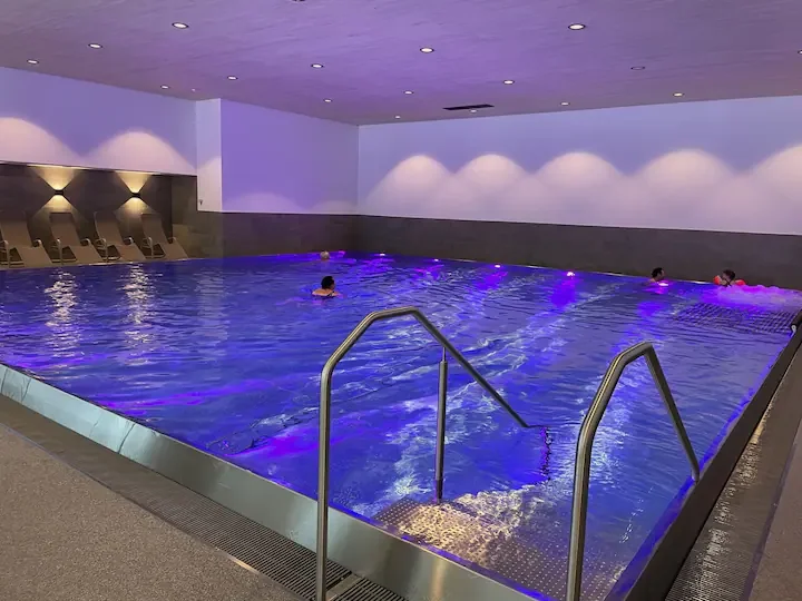 Indoor swimming pool with purple lighting, several people swimming, and lounge chairs along the wall.