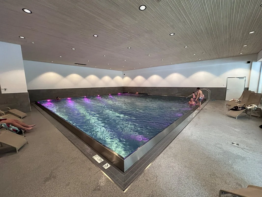 Indoor swimming pool with people relaxing and swimming, surrounded by lounge chairs and a modern ceiling with recessed lighting.