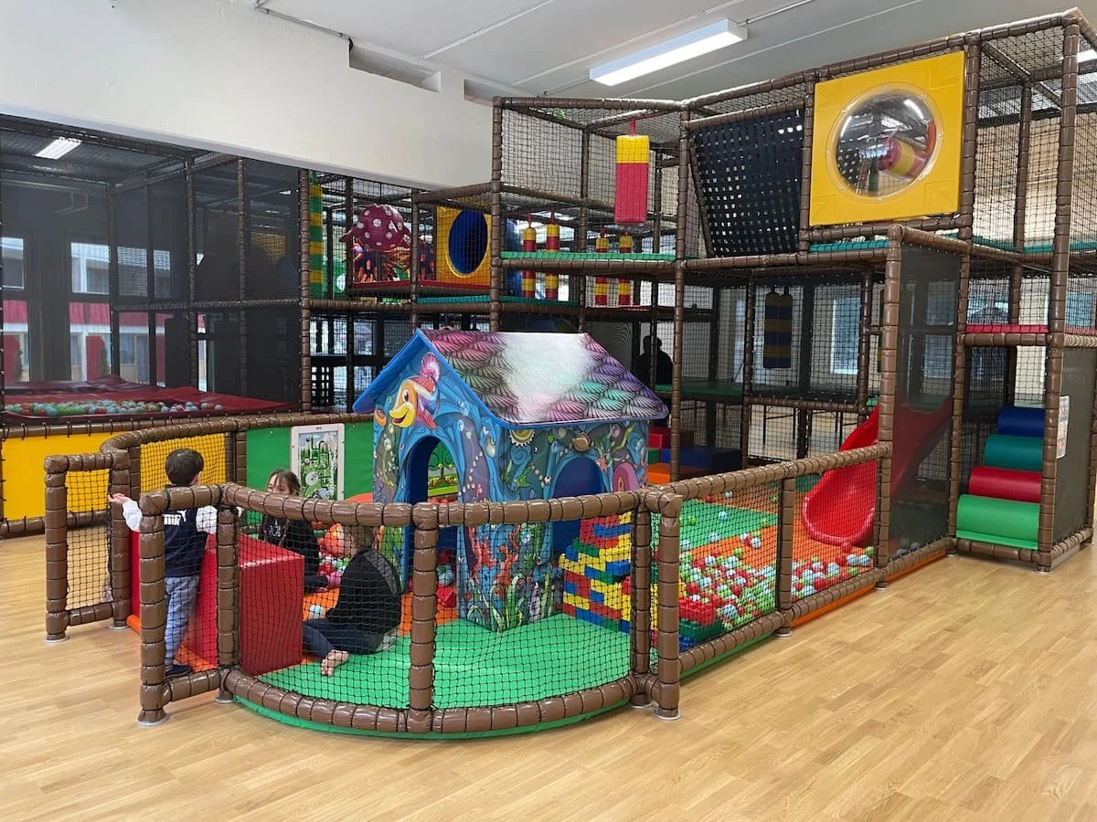 Indoor kids playground with various play structures, slides, ball pits, and soft climbing areas, surrounded by netting for safety, with children playing inside.